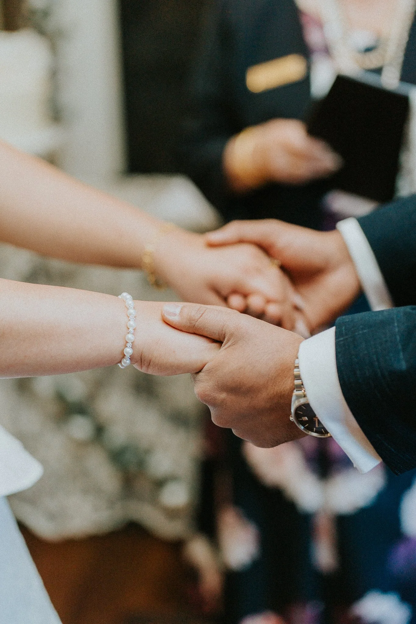 Close-up of two people holding hands during a wedding or formal ceremony, with a woman wearing a pearl bracelet and a man dressed in a dark suit with a white shirt and a watch.