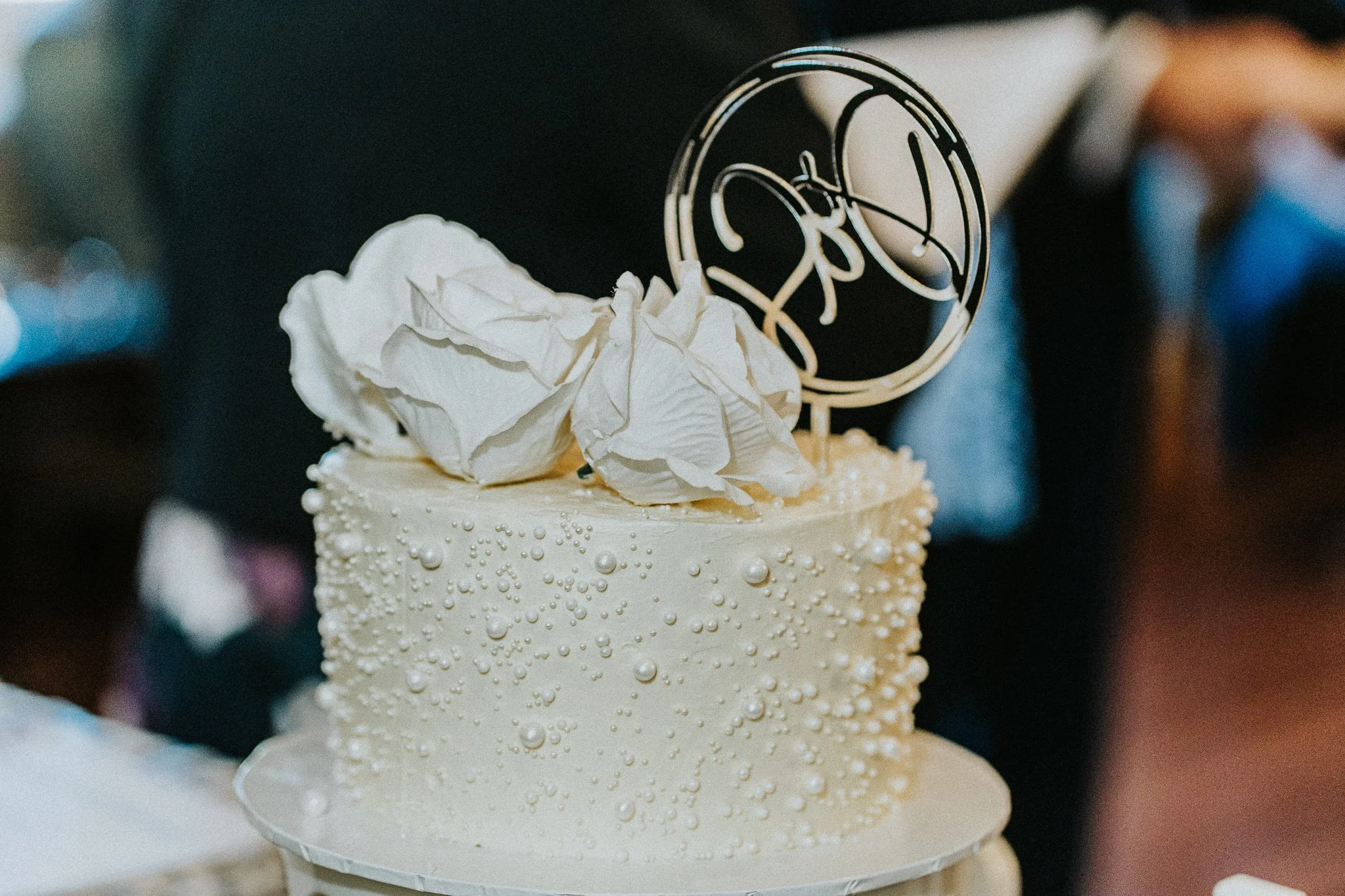 A decorated white cake with pearls, topped with crumpled white paper and a circular cake topper with white and gold design, possibly announcing a celebration like a wedding.