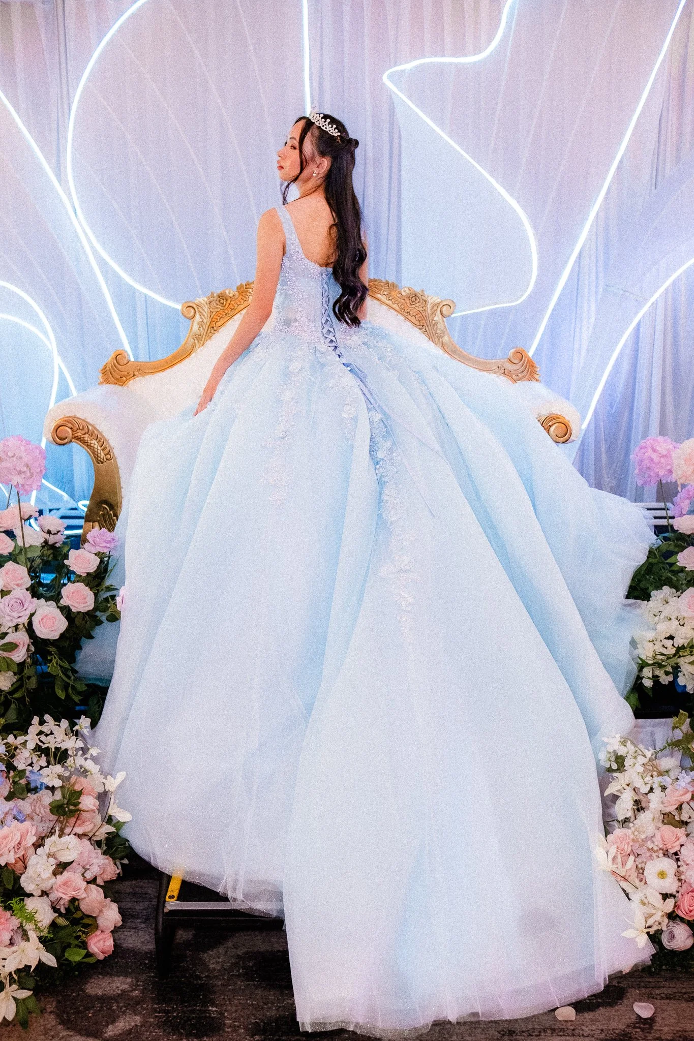 A woman in a light blue ball gown with lace details, wearing a tiara, sitting on a decorated sofa with pink flowers around.
