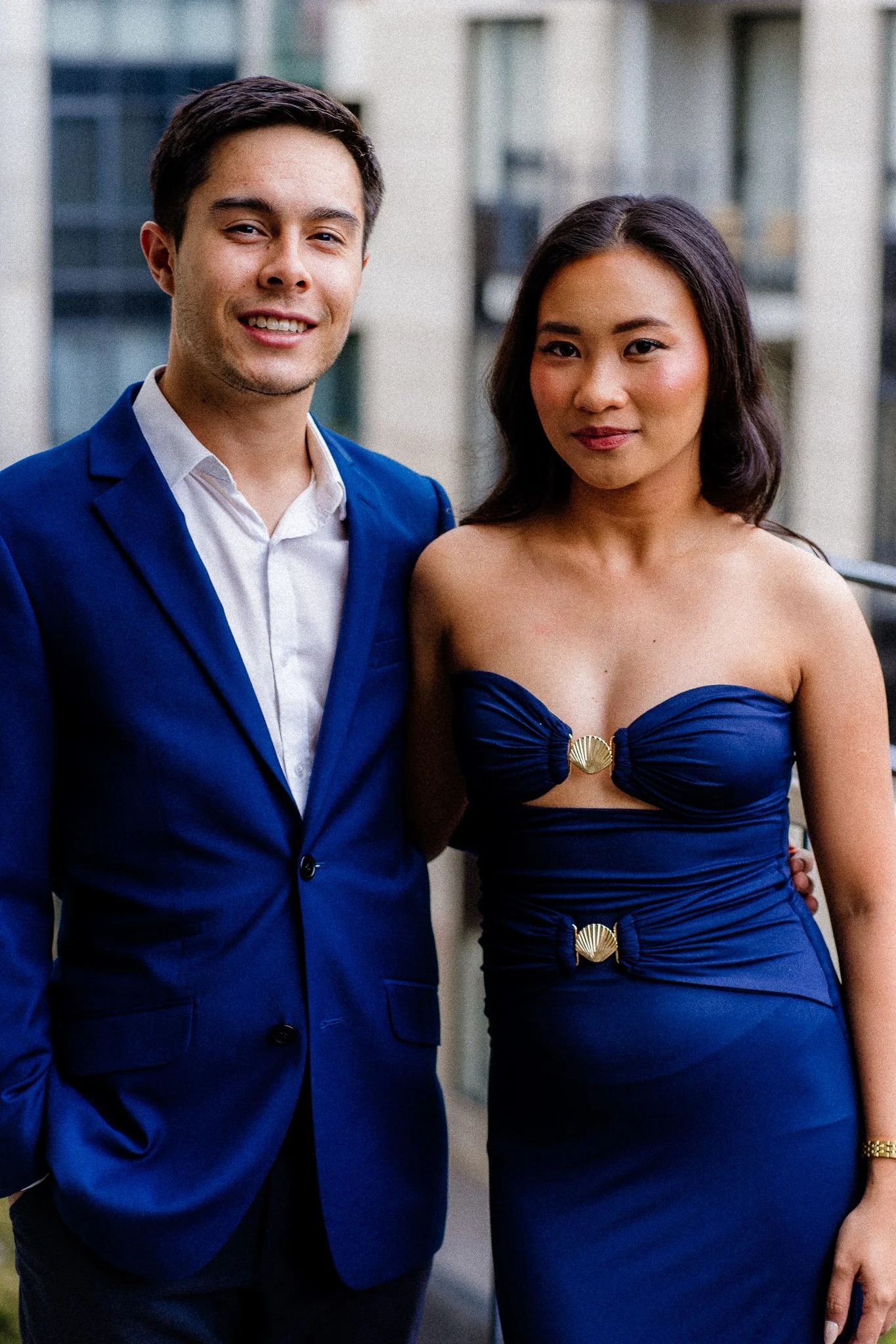 A man in a blue suit smiling and a woman in a strapless blue dress with shell-shaped embellishments on the front, standing on a balcony with modern buildings in the background.