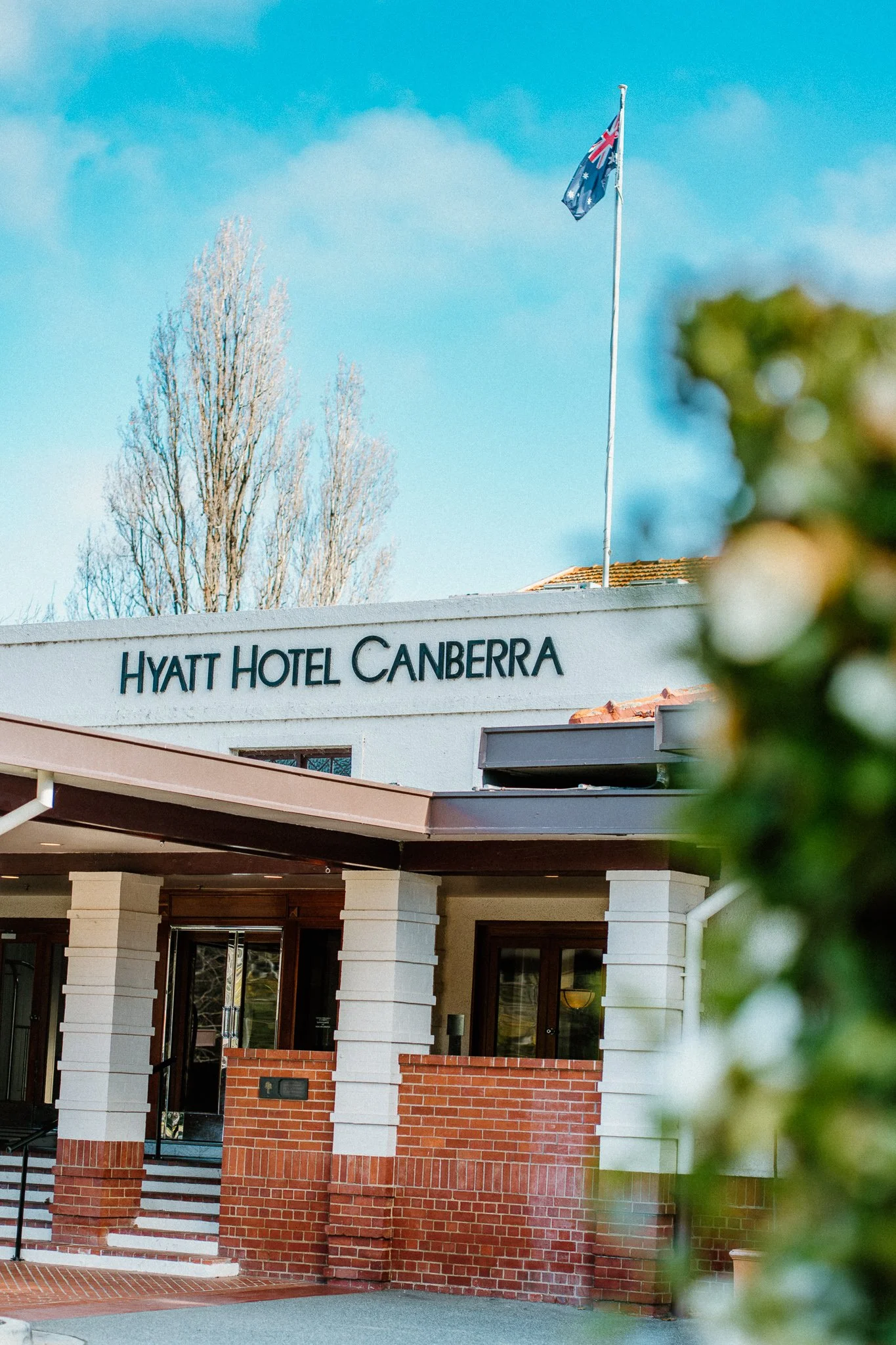 Exterior of the Hyatt Hotel Canberra with a sign displaying its name, a flagpole with the Australian flag, part of the hotel entrance, and a tall tree in the background.