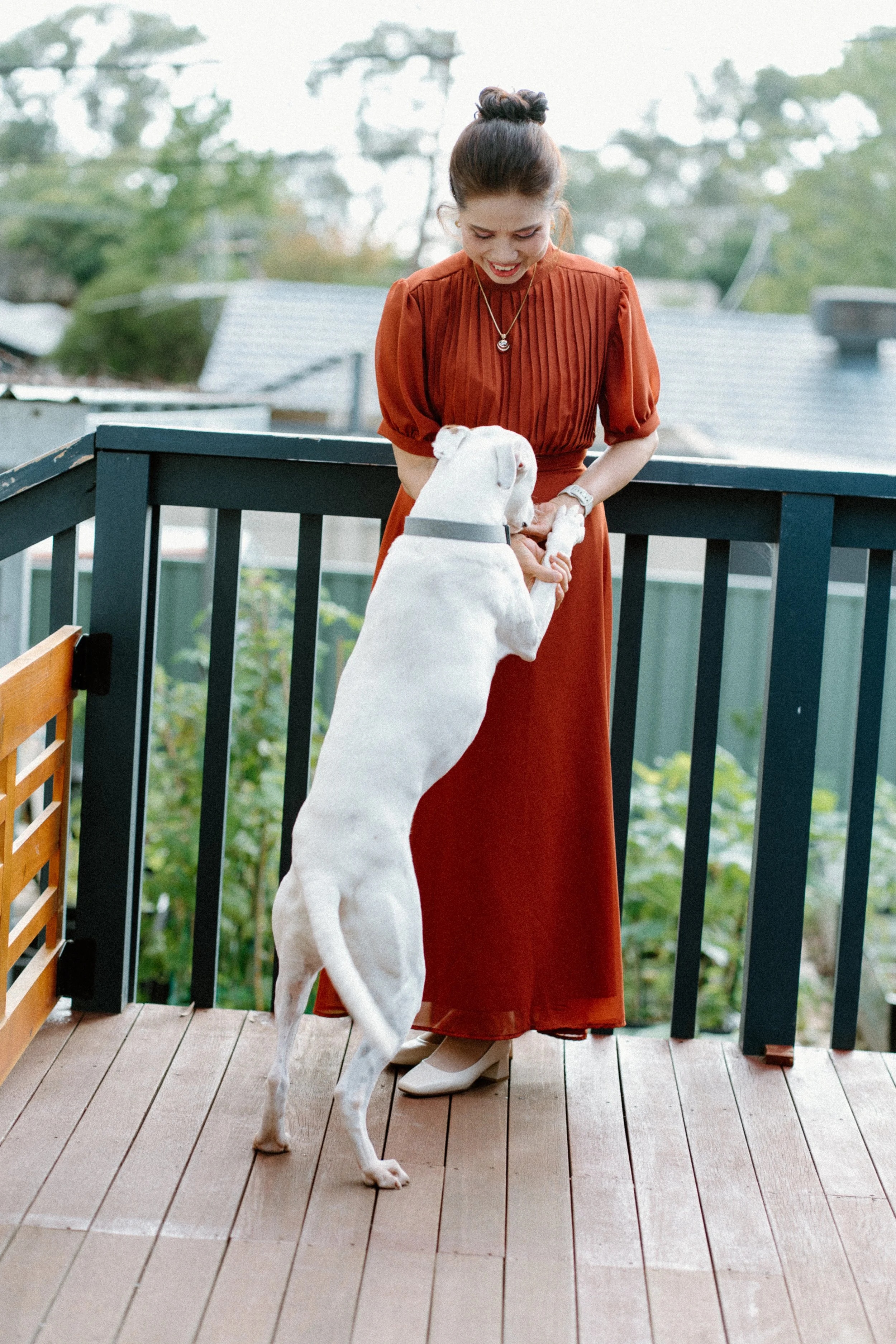 A woman in an orange dress holding a white dog on a deck outside.
