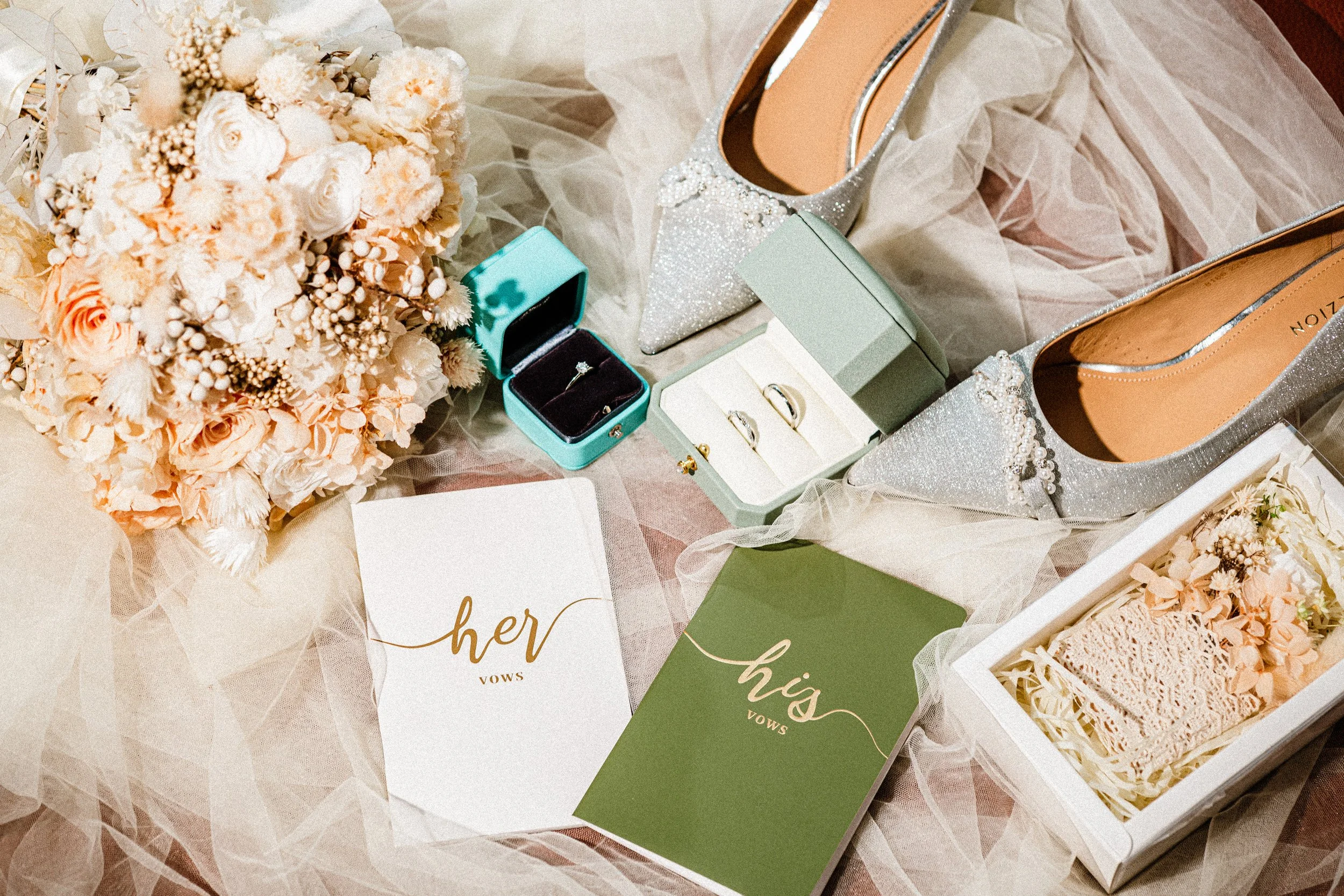 A collection of wedding accessories including a bouquet of cream and blush flowers, a pair of sparkly silver high heels with pearl embellishments, open jewelry boxes containing rings, and greeting cards with the words 'her vows' and 'his vows' writte