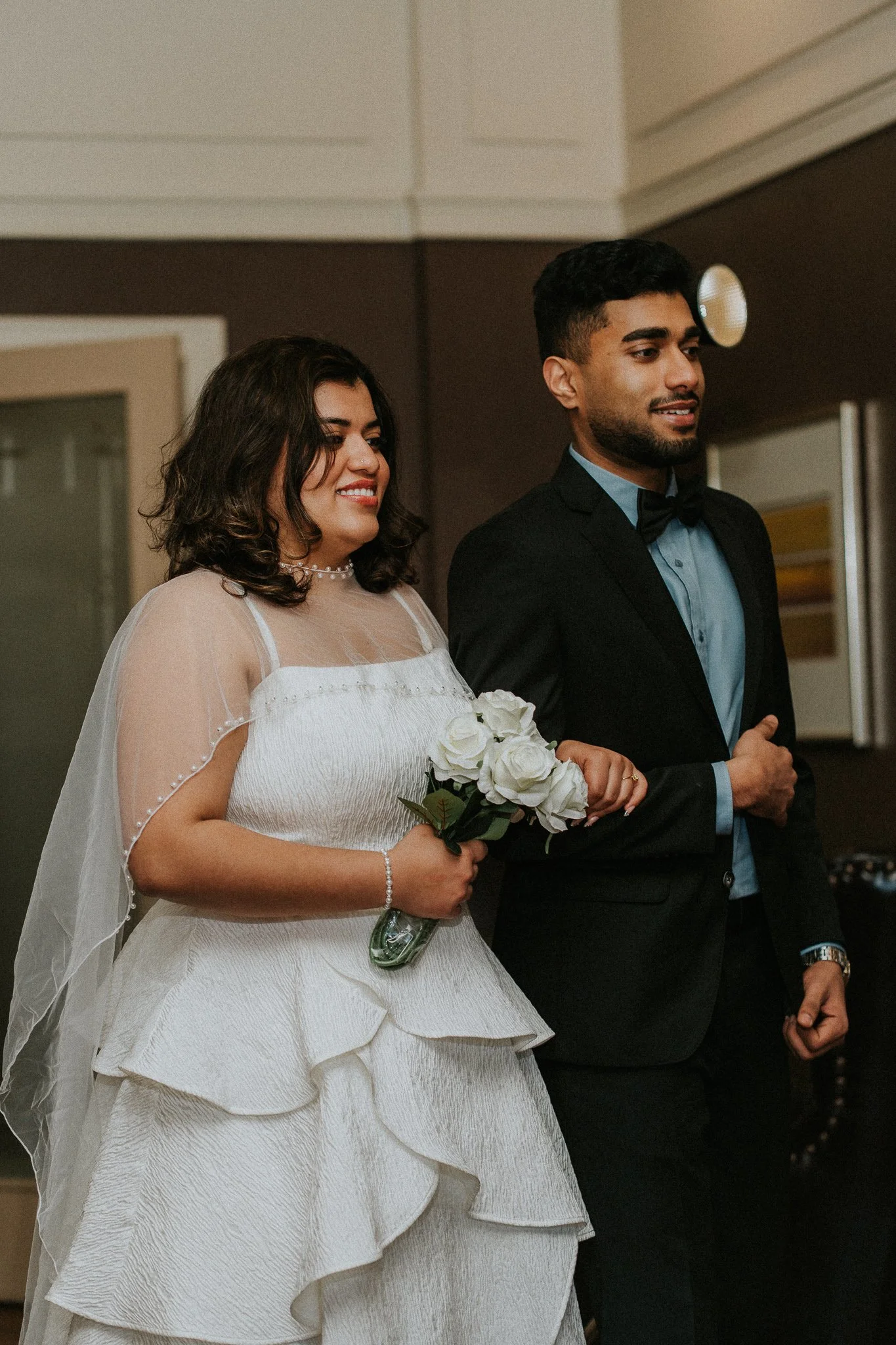 A bride and groom standing side by side during their wedding ceremony. The bride is holding a bouquet of white roses and dressed in a white gown with sheer sleeves, while the groom is wearing a black tuxedo with a light blue shirt and a black bow tie