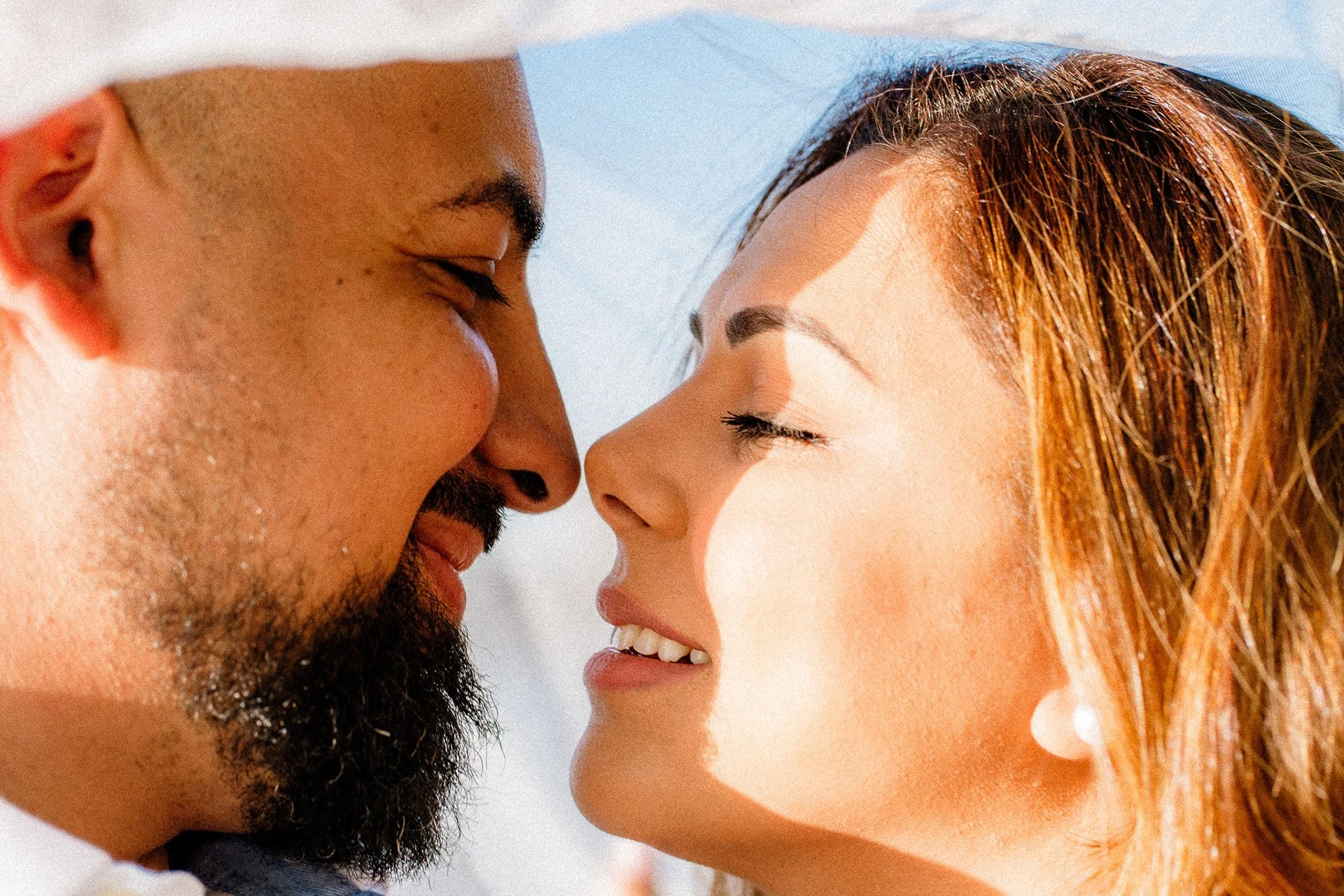 A man and a woman face each other with foreheads touching, smiling with their eyes closed against a clear blue sky.