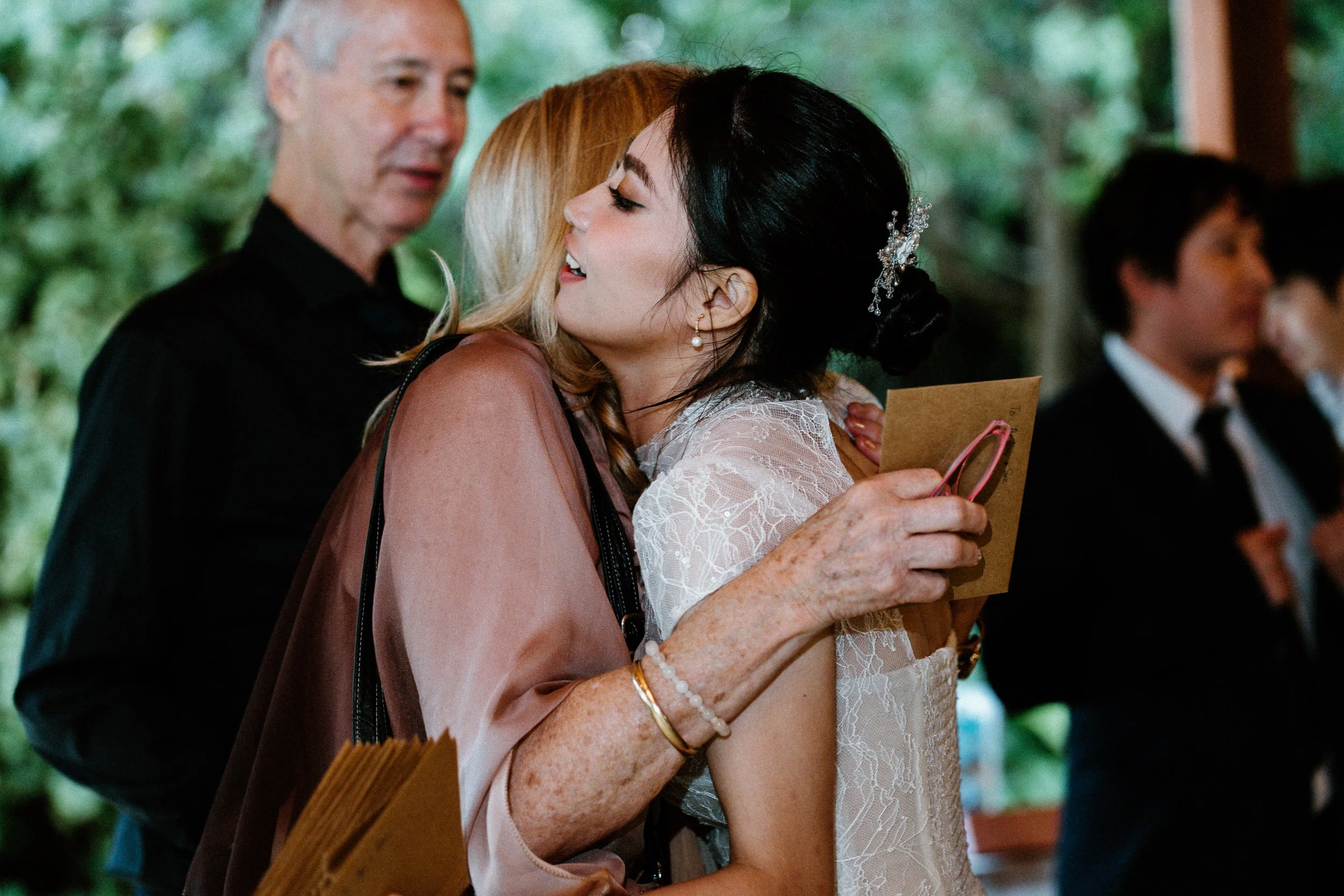 An elderly woman with white hair and freckles embraces a young woman with black hair and a decorative hairpiece at what appears to be a wedding or formal event.
