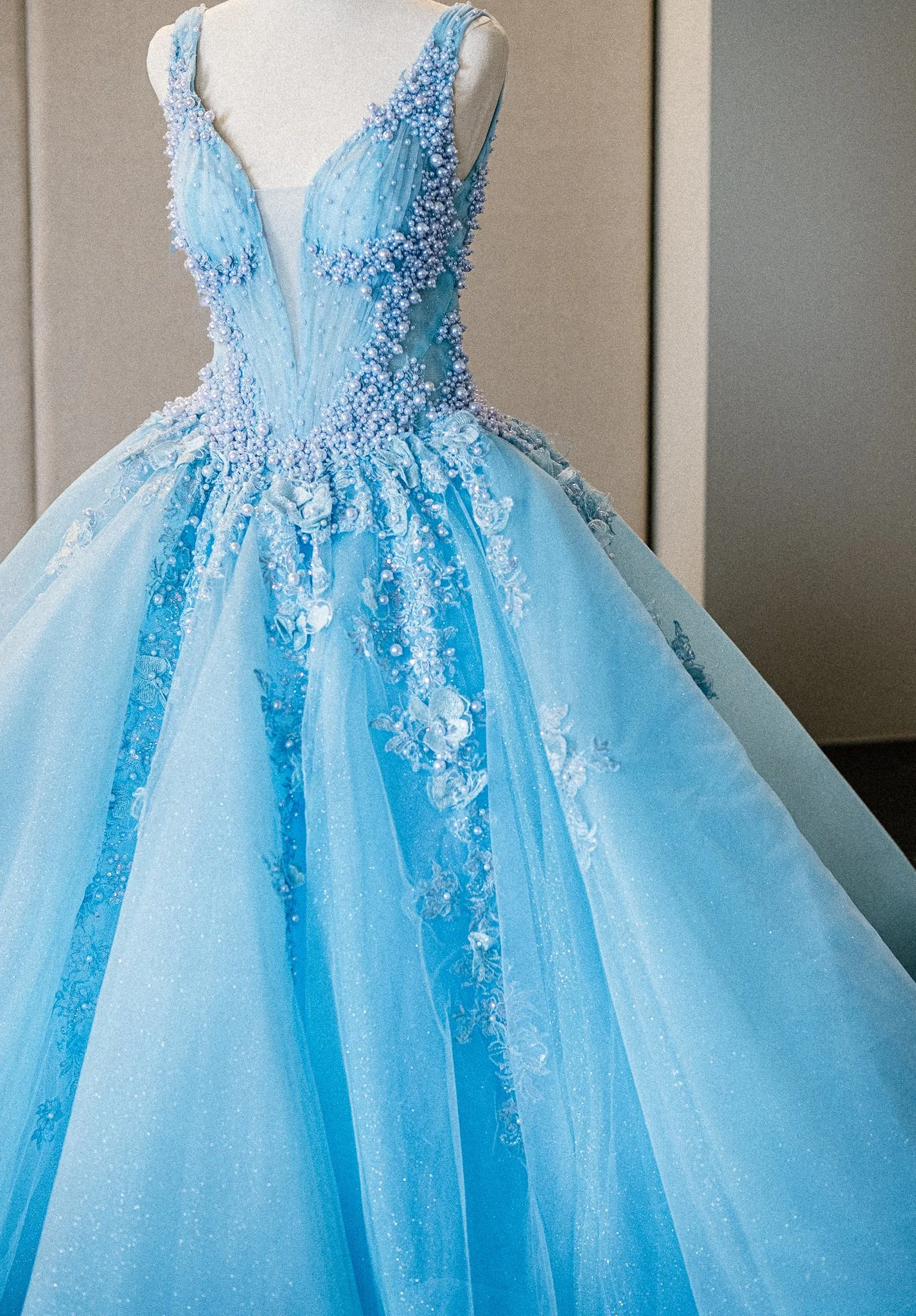 A light blue ball gown with elaborate pearl and floral embellishments on the bodice and straps, displayed on a mannequin.