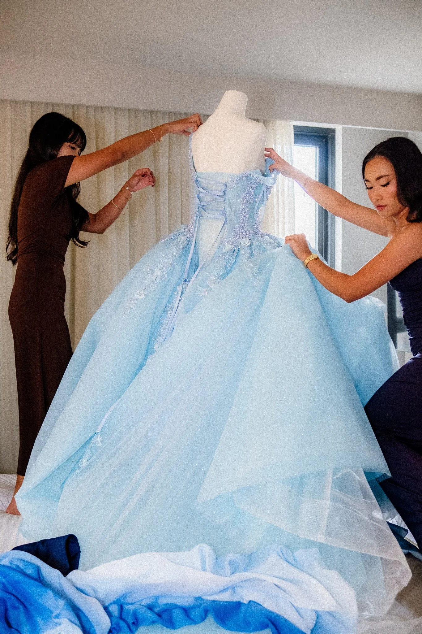Two women are adjusting a light blue ball gown with lace and pearl embellishments on a mannequin, in a room with natural light from a window.