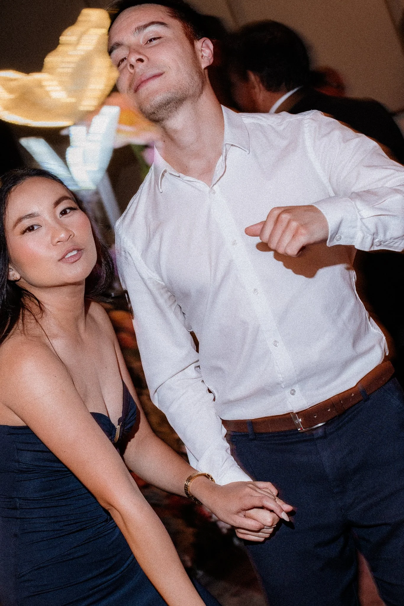 A man and woman holding hands at a social event or party, both dressed in formal attire. The woman is wearing a blue strapless dress and the man is in a white dress shirt with rolled-up sleeves, brown belt, and dark pants. The background shows other 