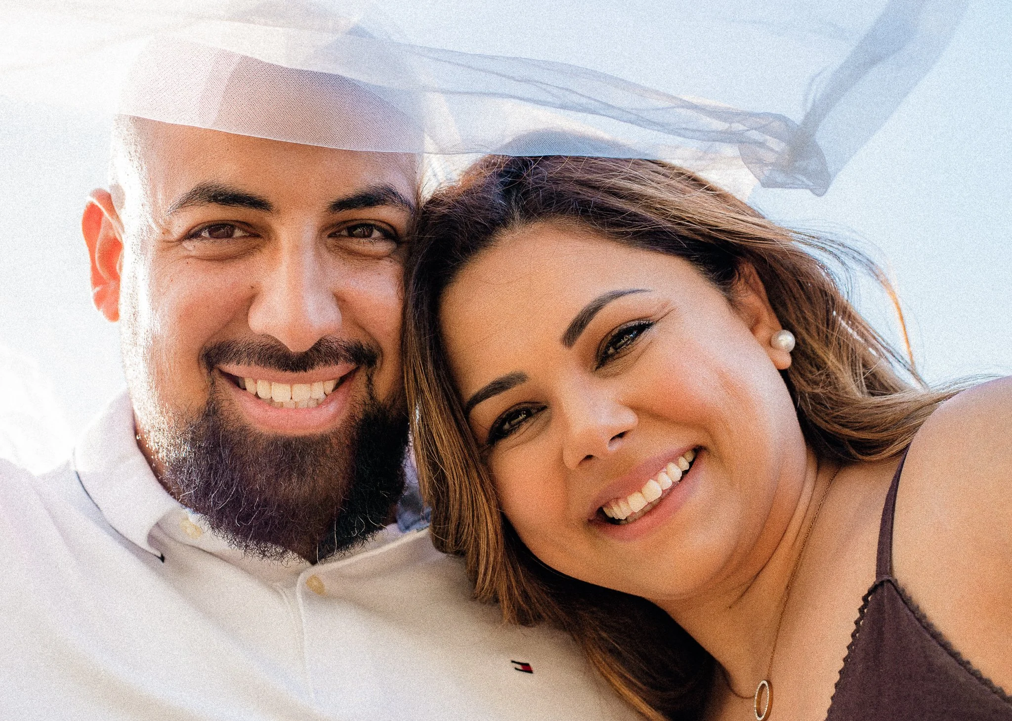 Close-up of a smiling man with a beard and mustache and a woman with long hair, both smiling and looking at the camera, with a light background.