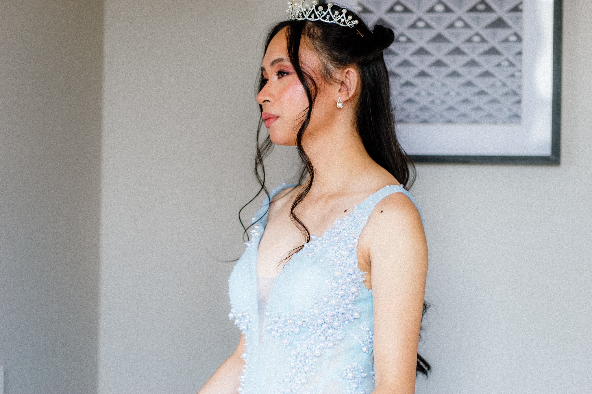A woman in a blue dress with pearl embellishments, wearing a tiara and pearl earrings, is seen in profile against a plain background with a framed artwork.