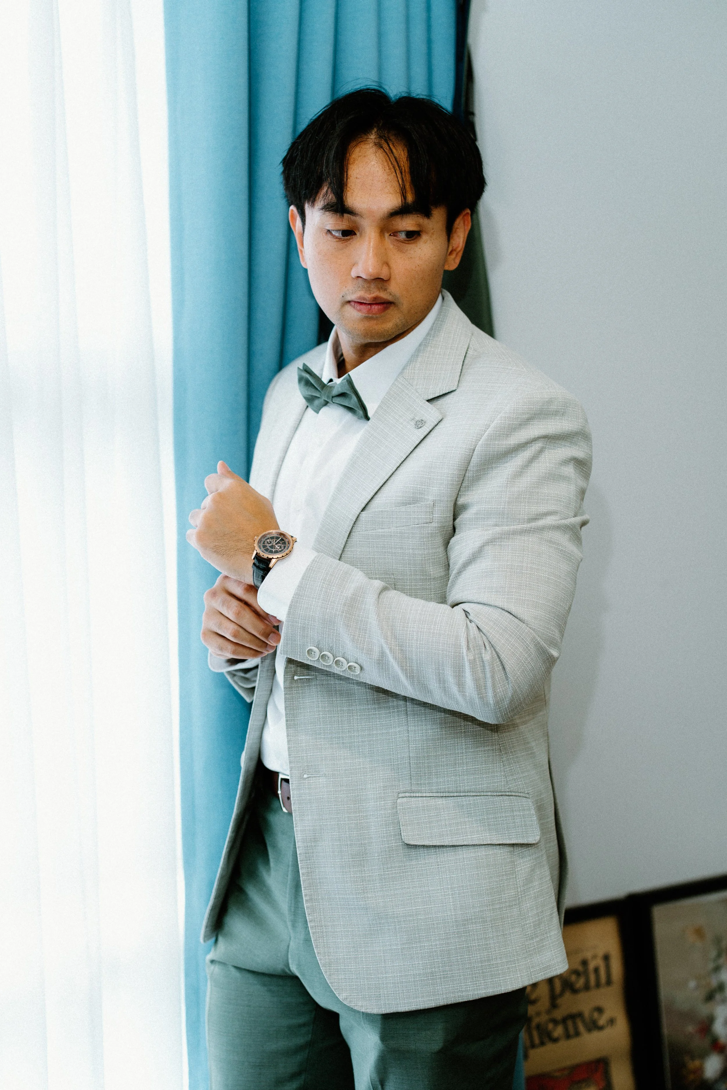 A young man with black hair, wearing a light gray suit, white shirt, and gray bow tie, looking down and adjusting his watch in front of blue curtains.