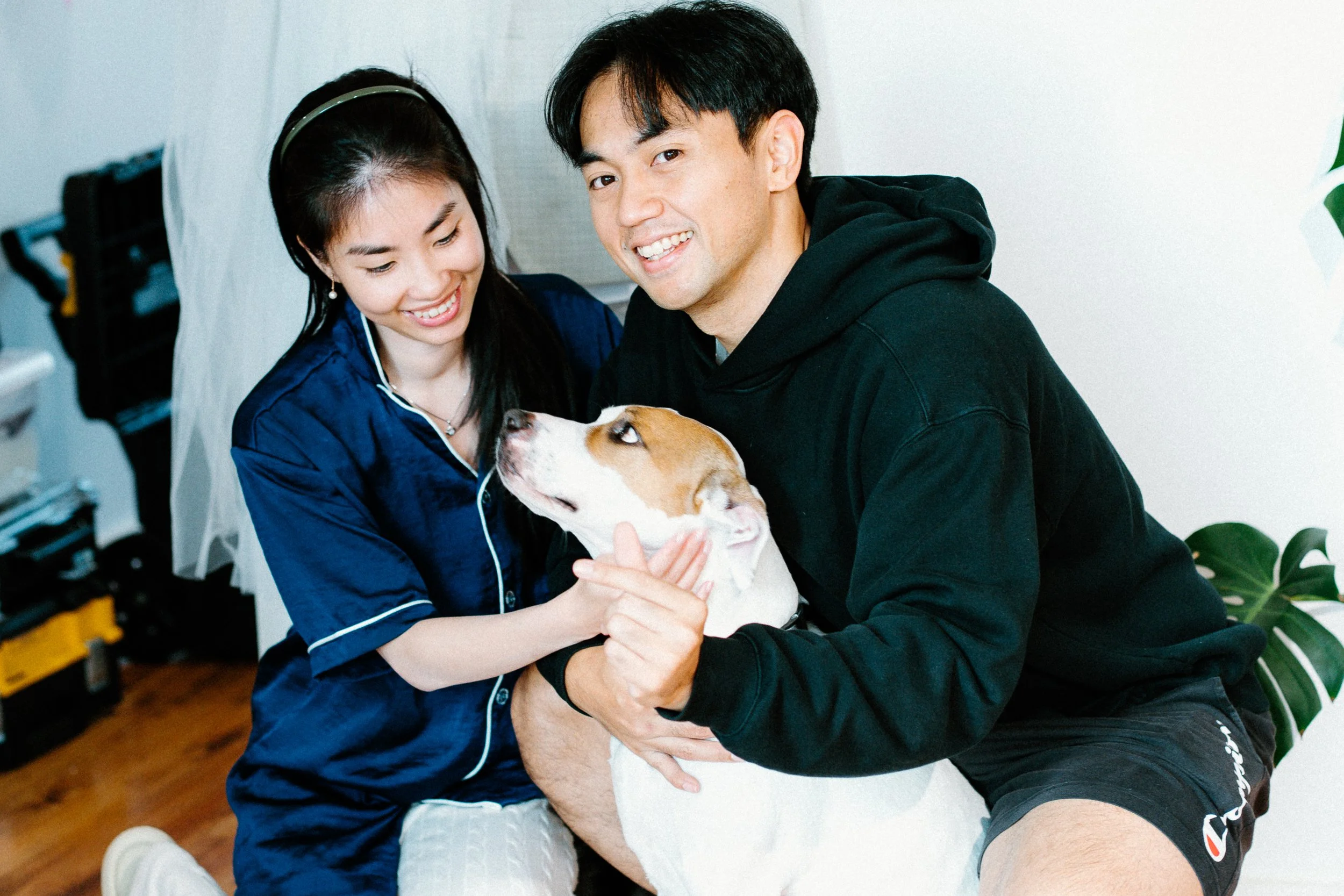 A young woman and a young man are smiling and holding a dog together indoors. The woman wears earrings and a blue shirt, while the man wears a black hoodie and shorts. The dog is white with brown patches, and they are all happy.