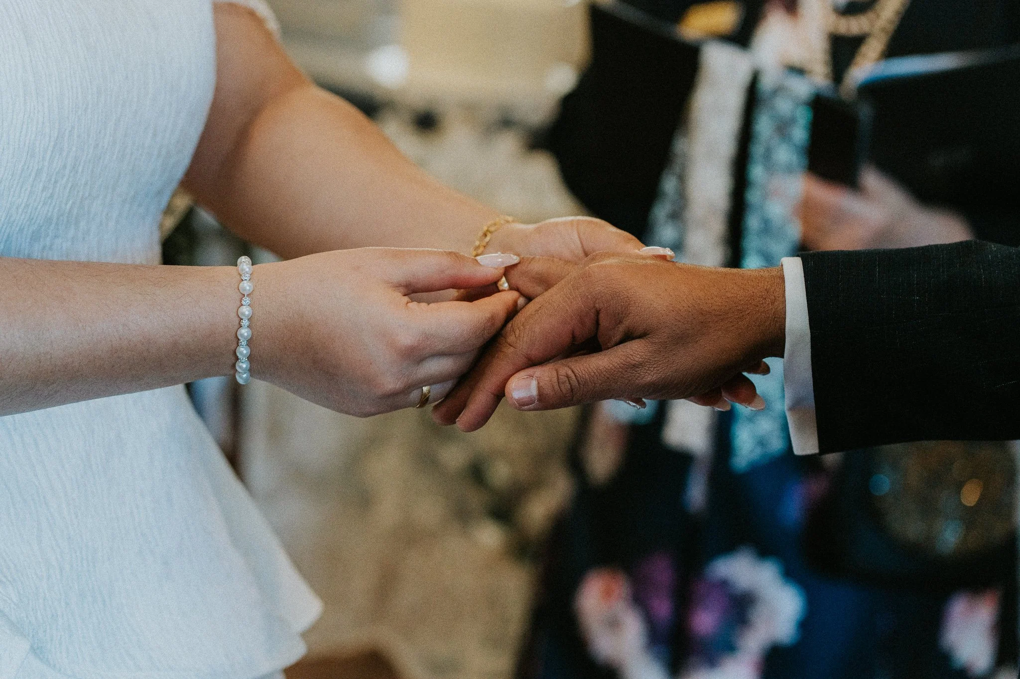 Close-up of a bride and groom holding hands during a wedding ceremony, with the bride wearing a pearl bracelet and wedding ring, and the groom wearing a tuxedo with a white shirt and black suit.