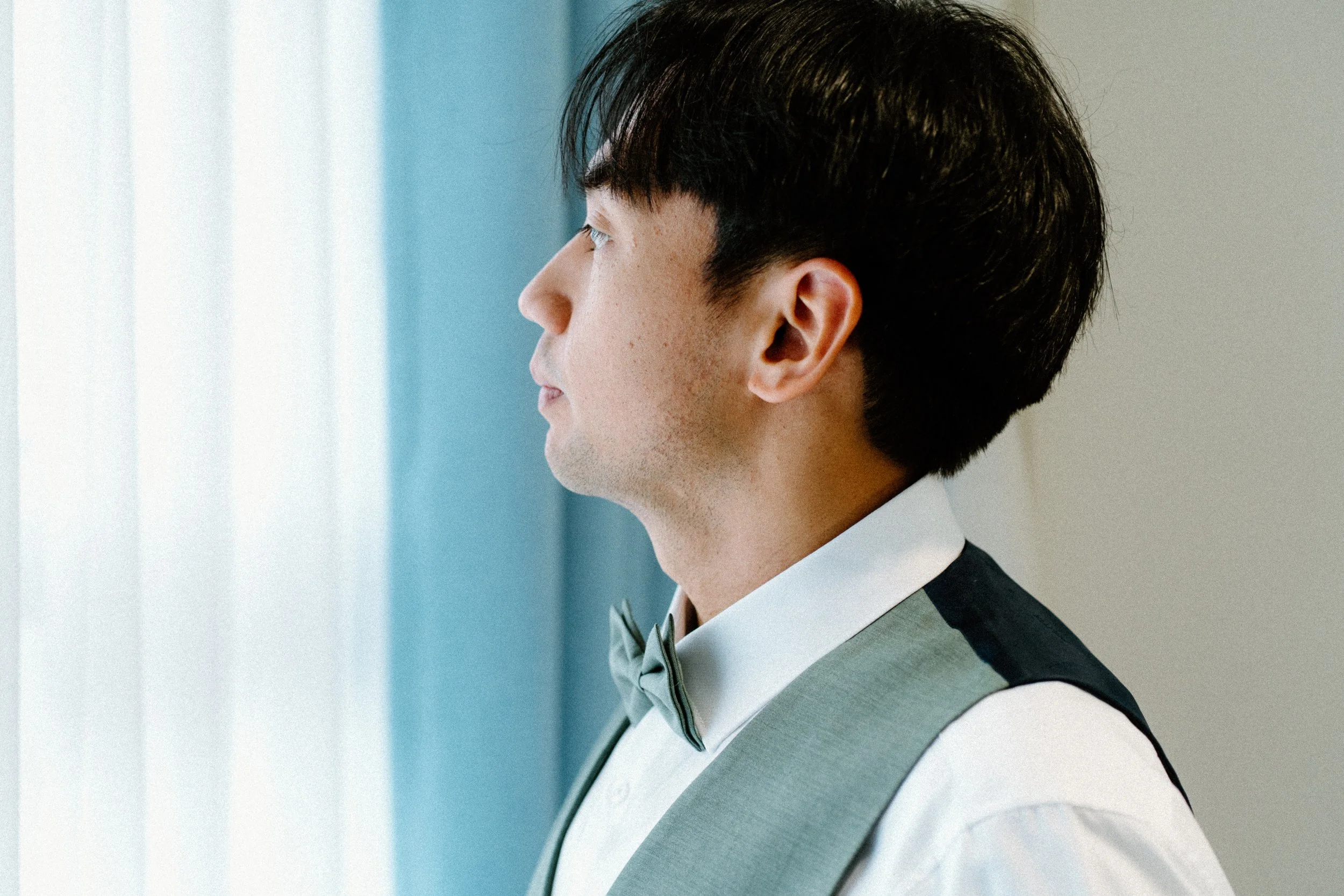Side profile of a young man dressed in a tuxedo with a bow tie, standing indoors near a window with blinds.