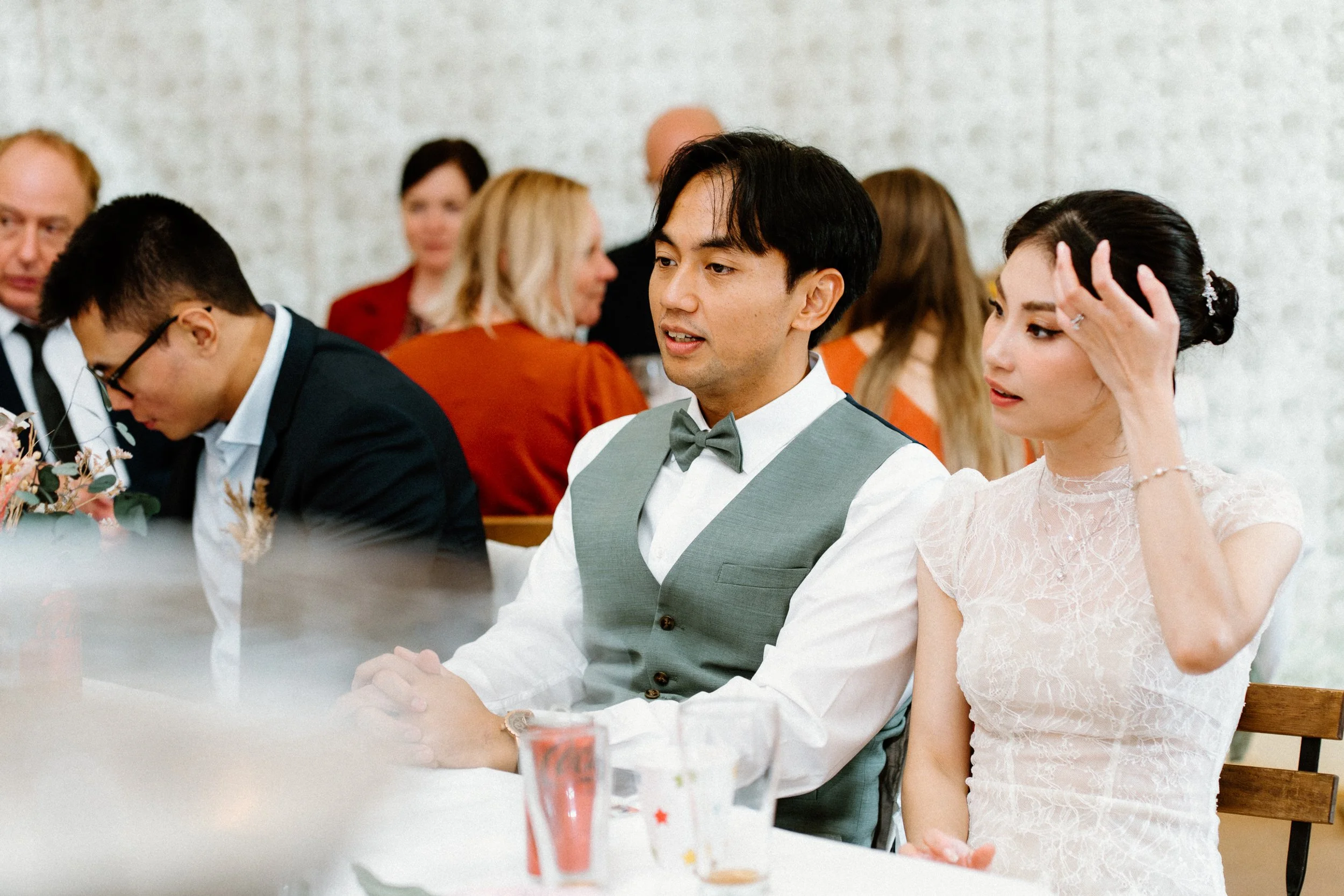 A group of people, including a man in a gray vest and bow tie and a woman in a white lace dress, sitting at a table during a social gathering or celebration.