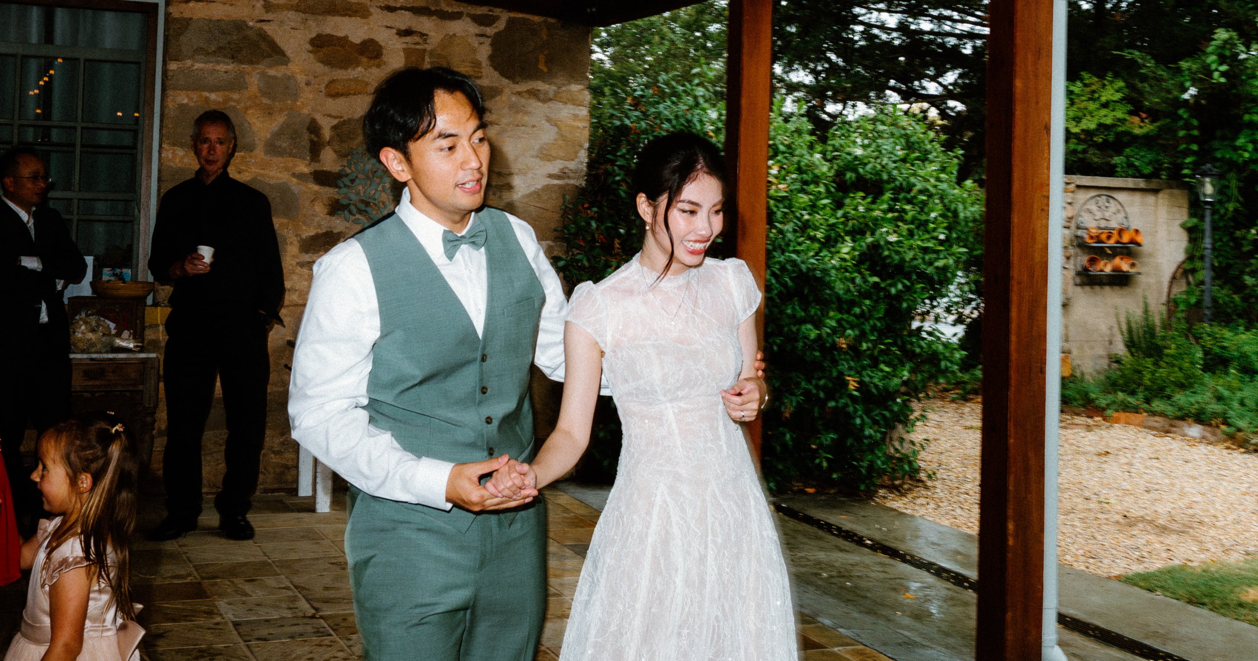 A couple dressed in wedding attire holding hands during their wedding celebration. The groom wears a grey vest and bow tie, and the bride wears a white lace dress. They are indoors with other guests and greenery visible outside.