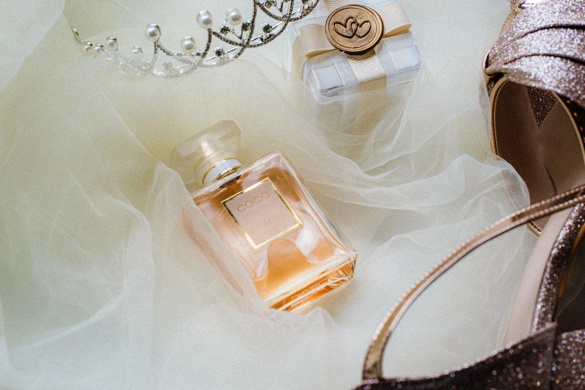 A gift wrapped with a beige ribbon and seal, a bottle of perfume labeled Coco, a satin red clutch, and a glittery pink shoe with sequins, all on white fabric.