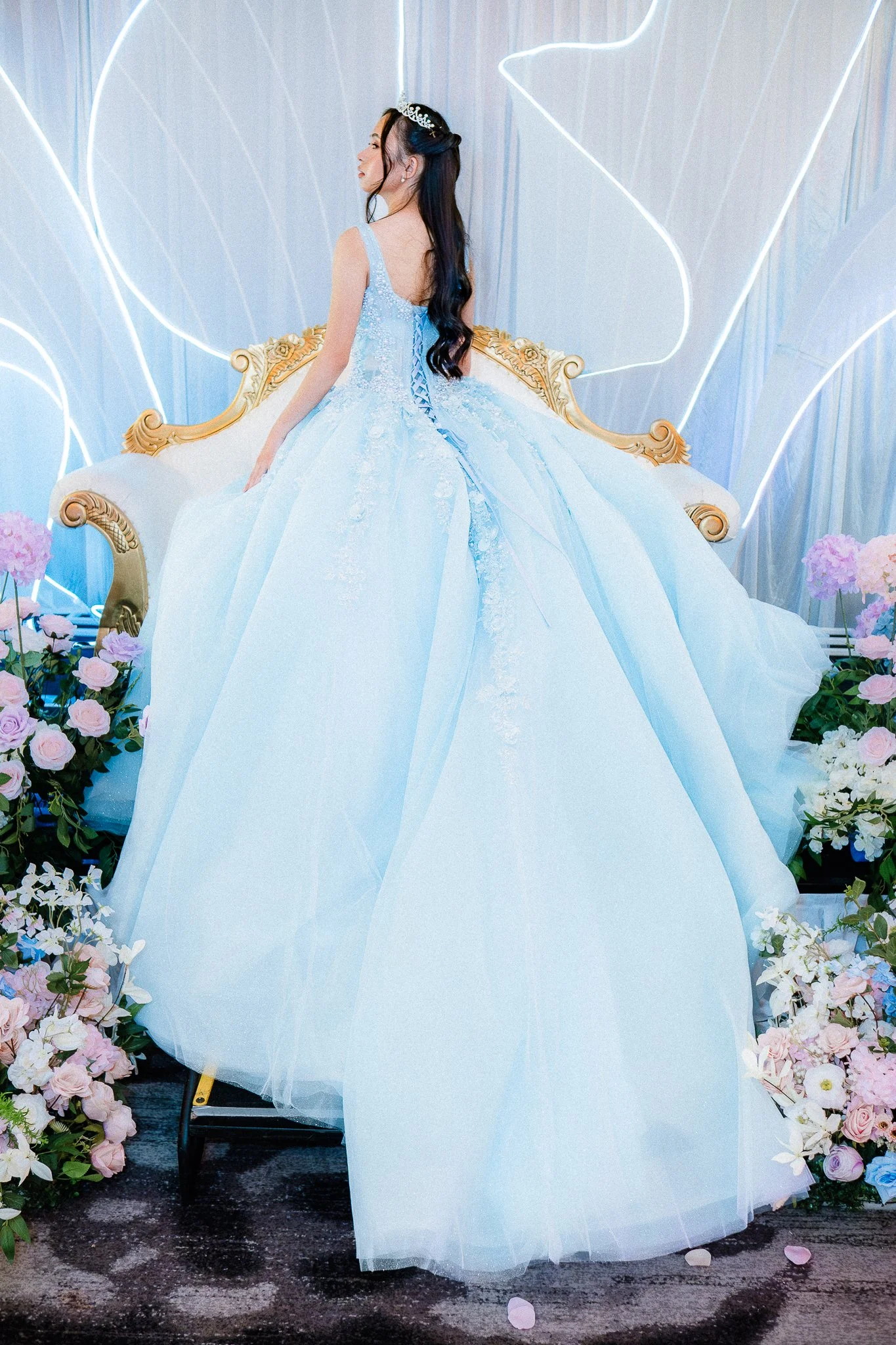A young woman wearing a light blue ball gown with lace and floral details, sitting on a vintage gold and white sofa, surrounded by pink and white flowers with a decorative backdrop of blue drapes and white neon lights.