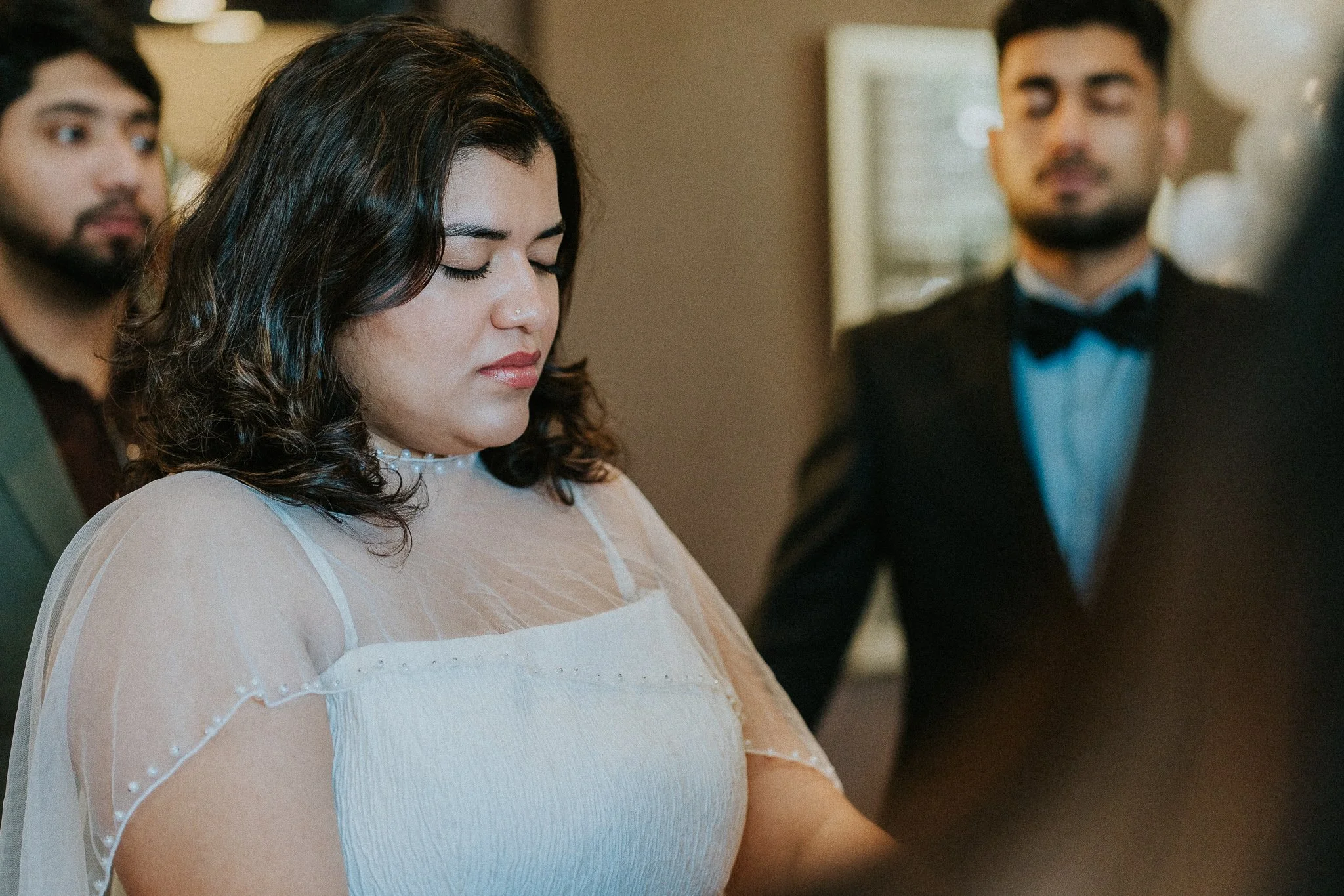 A woman with dark, curly hair and a nose piercing, dressed in white, standing with her eyes closed at a formal event. Two men in tuxedos are in the background, one looking at her and the other slightly out of focus.