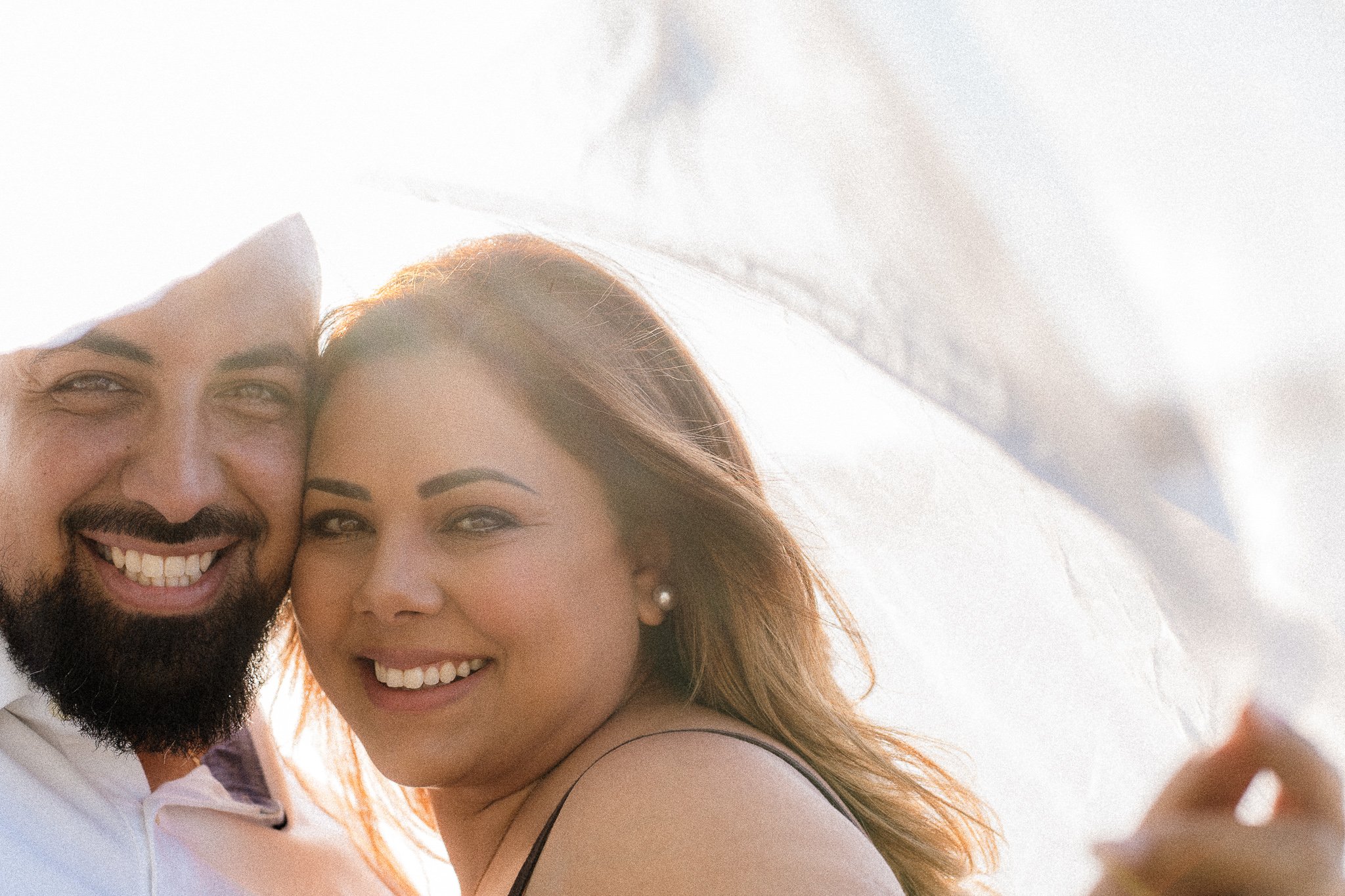 Close-up of a happy couple smiling, with the man having dark hair, a beard, and wearing a white shirt, and the woman having long reddish hair and wearing a black spaghetti strap top. They are outdoors in bright lighting.