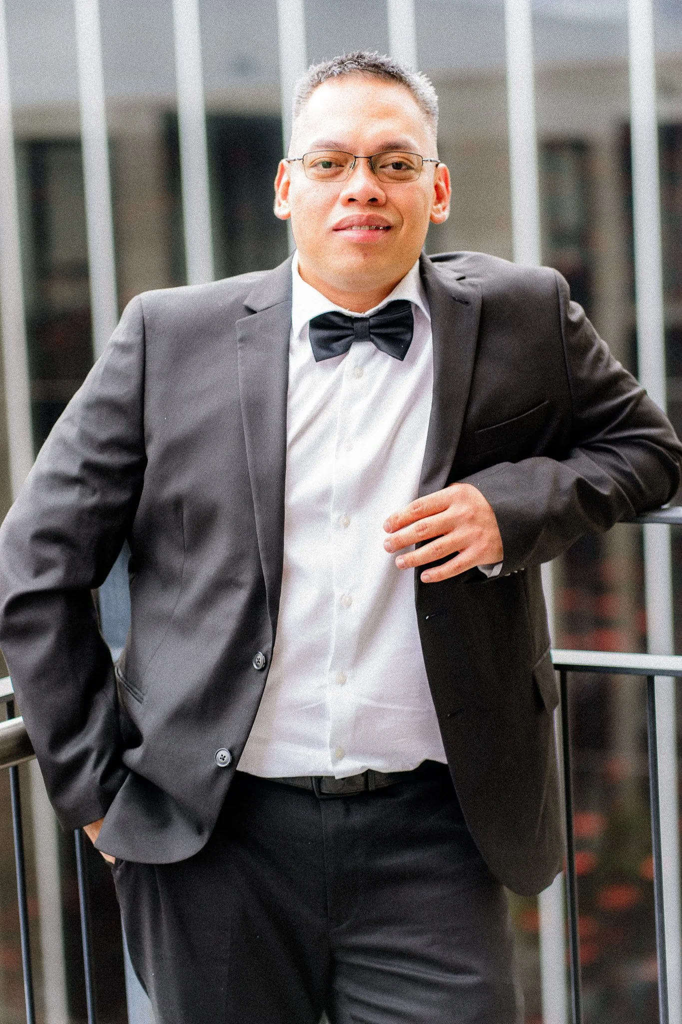 A man in a black tuxedo with a bow tie, white shirt, and glasses, standing outdoors near railing, smiling and looking at the camera.