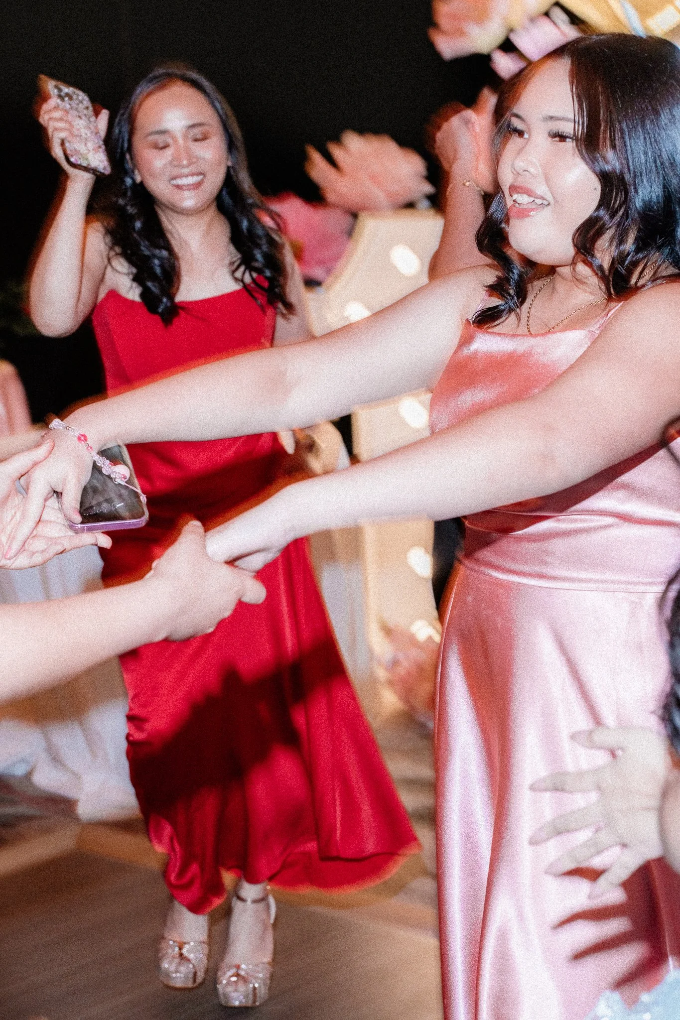 Two women are at a celebration, both wearing elegant dresses. One woman, in a pink satin dress, is smiling and looking at her friend. The other, in a red dress, also smiling with closed eyes, is holding a phone to take a photo. They are exchanging a 