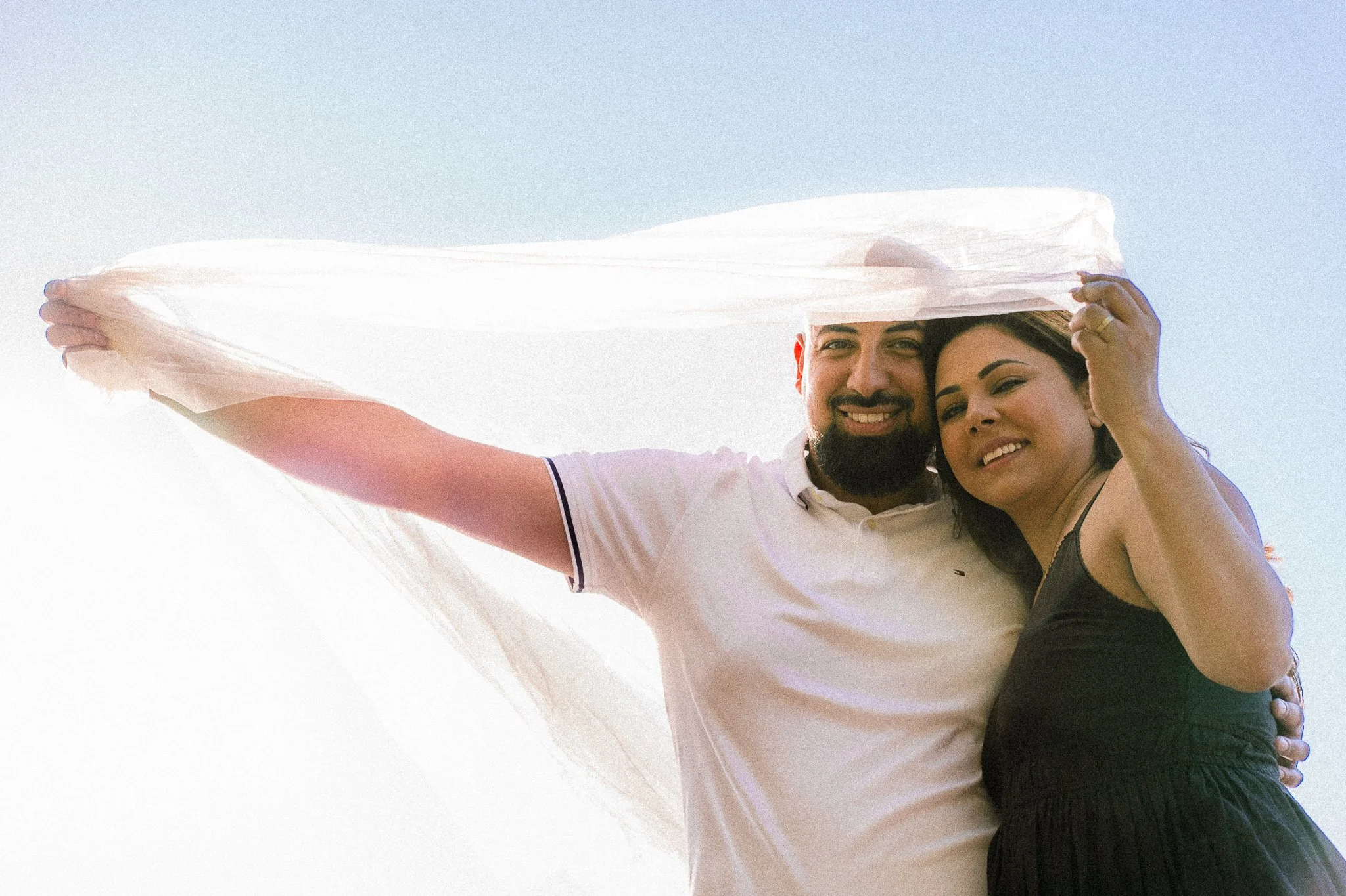 A happy couple taking a selfie together outdoors with a blue sky in the background, the woman is wearing a black strapless dress, and the man is wearing a white polo shirt.
