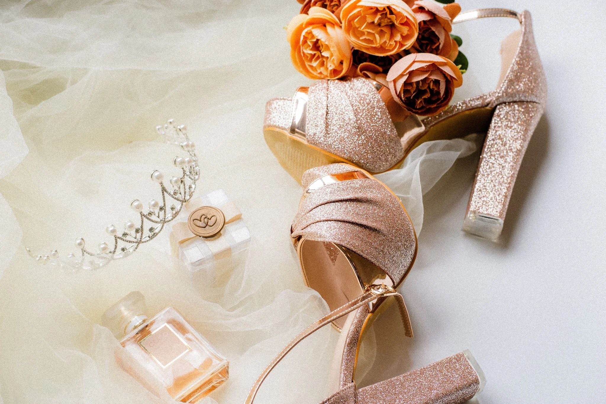 A collection of rose gold glitter high heel shoes, a bouquet of orange and peach flowers, a pearl headpiece, a perfume bottle, and a small box with a heart-shaped design on the lid, all laid out on a white surface.