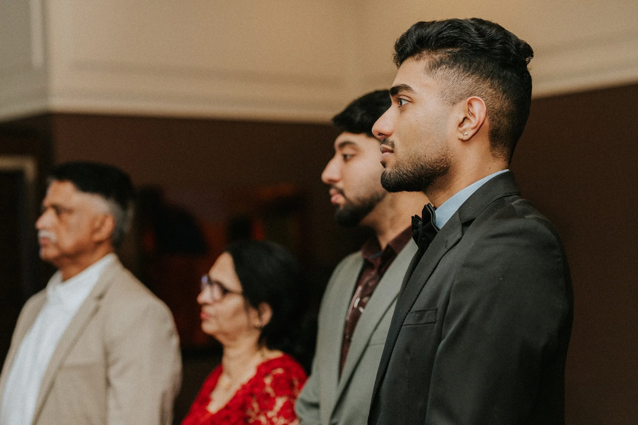 A group of diverse people dressed formally, standing in a line at a formal event, paying attention to something.