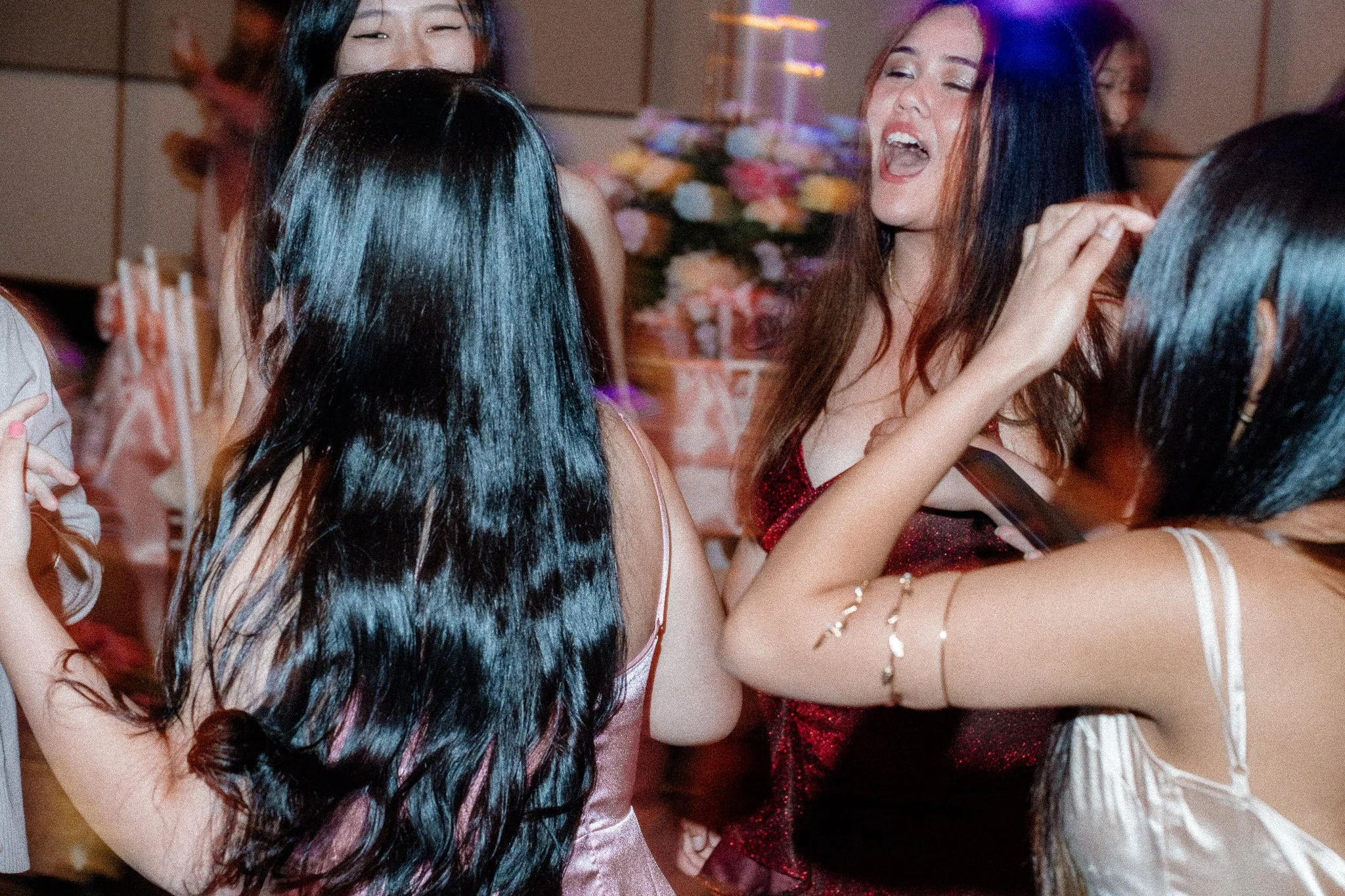 Group of women at a celebration or party, with some laughing and embracing, a floral arrangement in the background, wearing stylish dresses and jewelry.