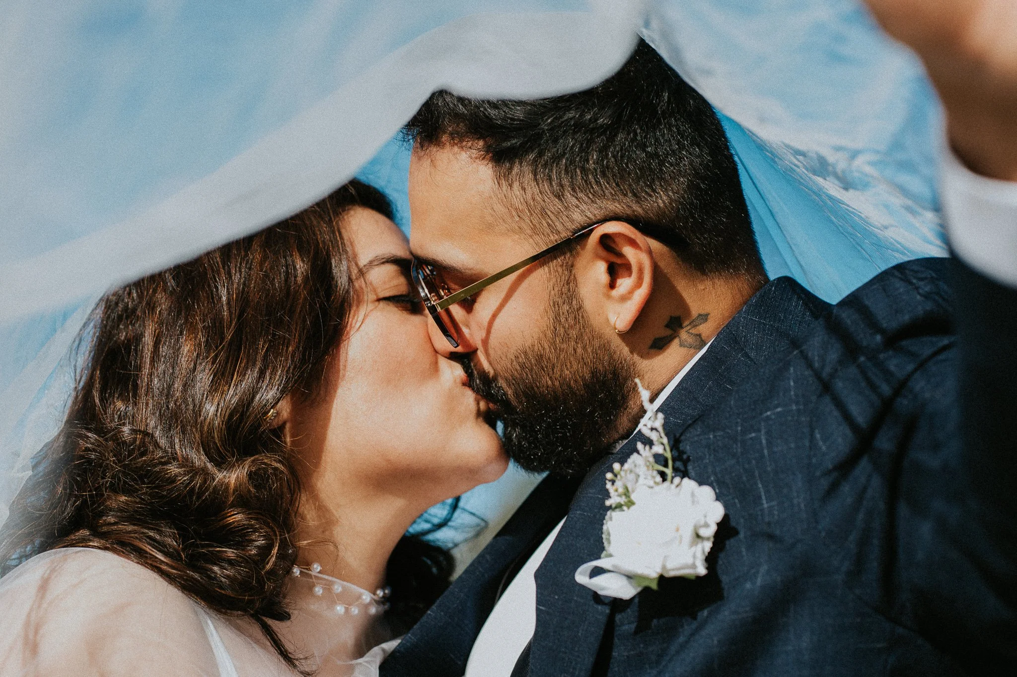 A couple kissing, the woman has long curly hair and is wearing a light dress, the man has dark hair, glasses, a tattoo on his neck, and is dressed in a dark suit with a white flower boutonniere.