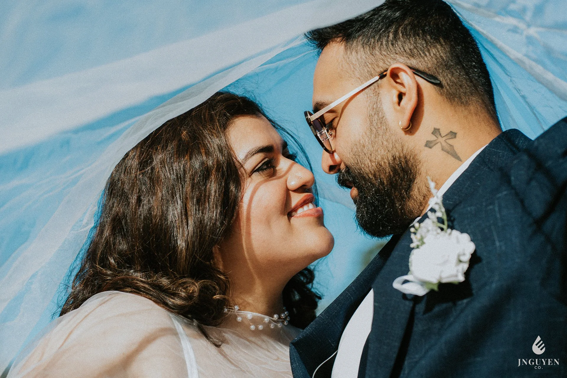 A bride and groom lying close together with their foreheads touching underneath a sheer veil, smiling and gazing into each other's eyes on a clear blue sky background.
