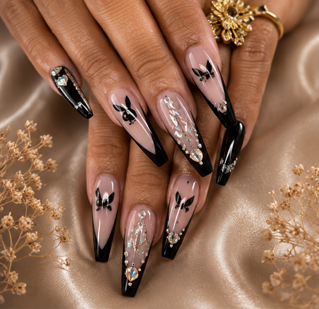 Close-up of a woman's hands displaying long, pointed acrylic nails with black French tips, bunny face designs, rhinestones, and iridescent accents, on a satin fabric background with gold floral decorations.