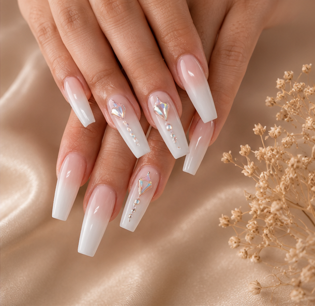 Close-up of a hand with long, white ombre nails decorated with rhinestones and small pearls, set against a satin background with dried flowers.