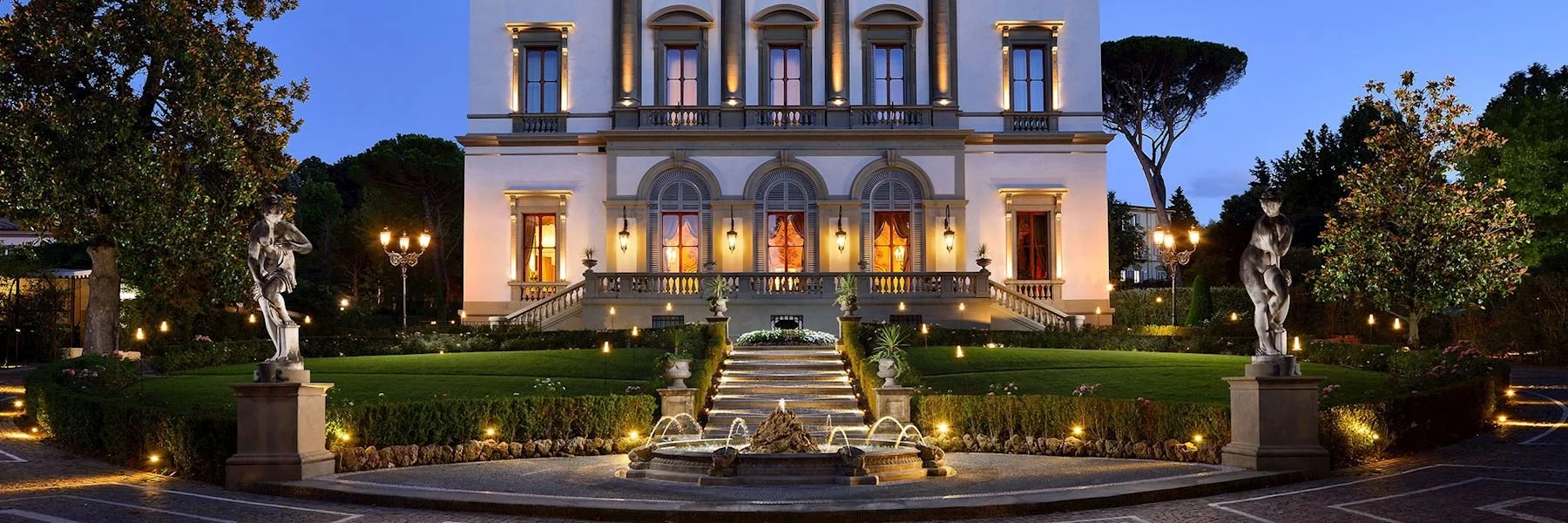 Elegant mansion illuminated at night with a garden, fountain, sculptures, trees, and decorative lighting.