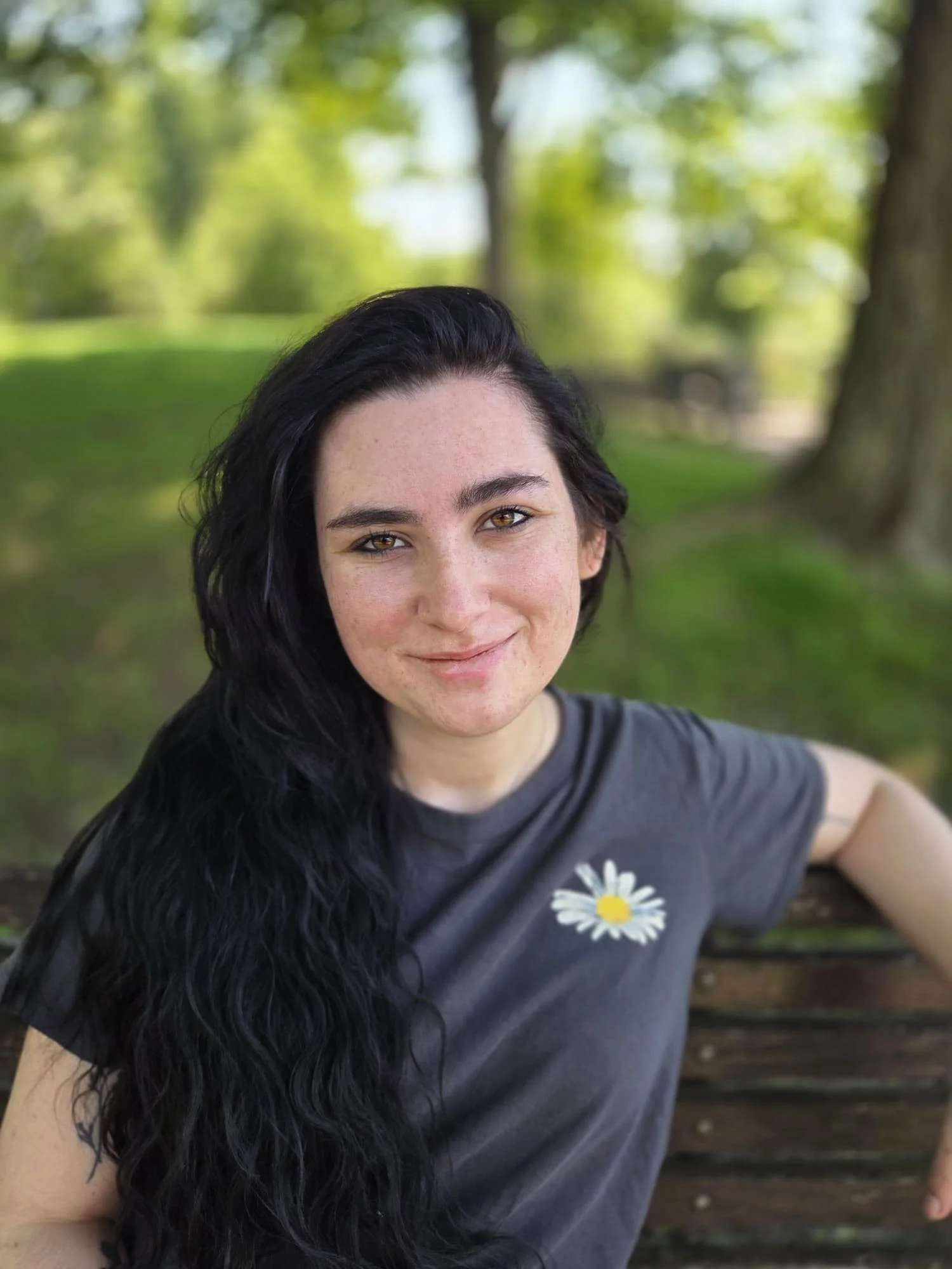 A woman with long dark wavy hair and light skin smiling outdoors, sitting on a wooden bench with a blurred green background of trees and grass, wearing a gray t-shirt with a white daisy design.
