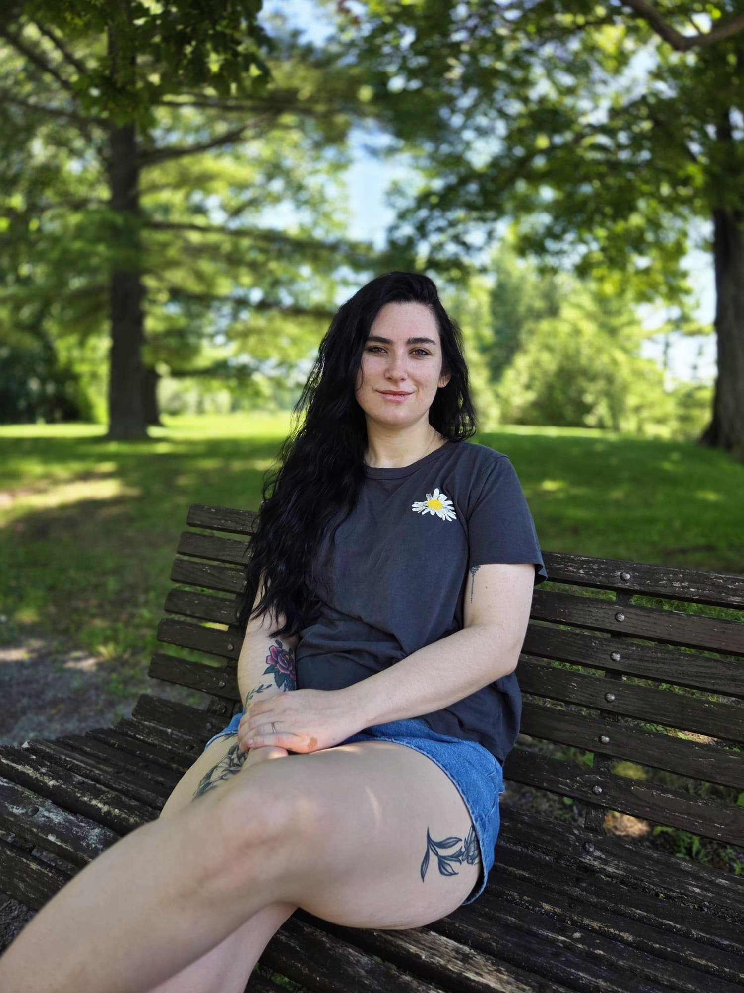 A young woman with long dark hair and tattoos on her arm sitting on a wooden park bench, with a background of lush green trees and grass, smiling at the camera.