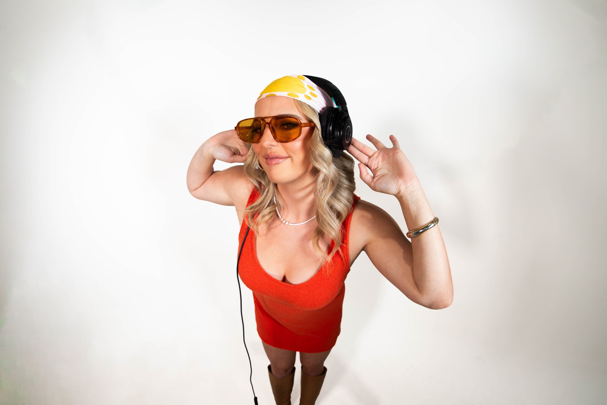 Woman with blonde hair wearing large orange sunglasses, a white headscarf with yellow polka dots, red sleeveless top, headphones, and tan knee-high boots, standing against a white background.