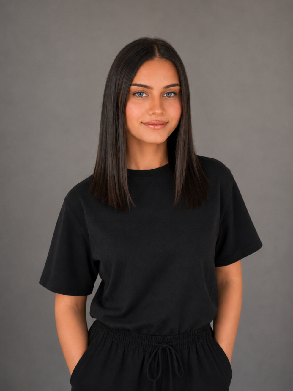 A young woman with shoulder-length dark brown hair, blue eyes, and light skin, wearing a black T-shirt and black sweatpants, standing against a gray background.