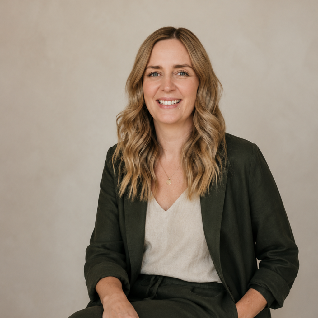A woman with wavy blonde hair, wearing a dark blazer and light-colored blouse, smiling at the camera against a neutral background.