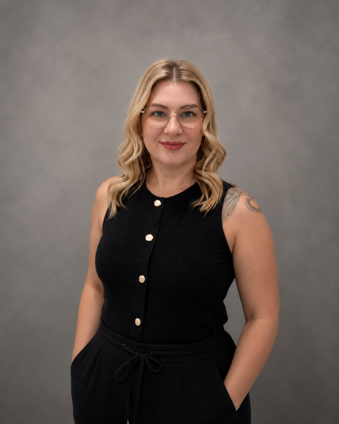 A woman with blonde wavy hair, glasses, wearing a sleeveless black top with gold buttons, standing against a gray background.