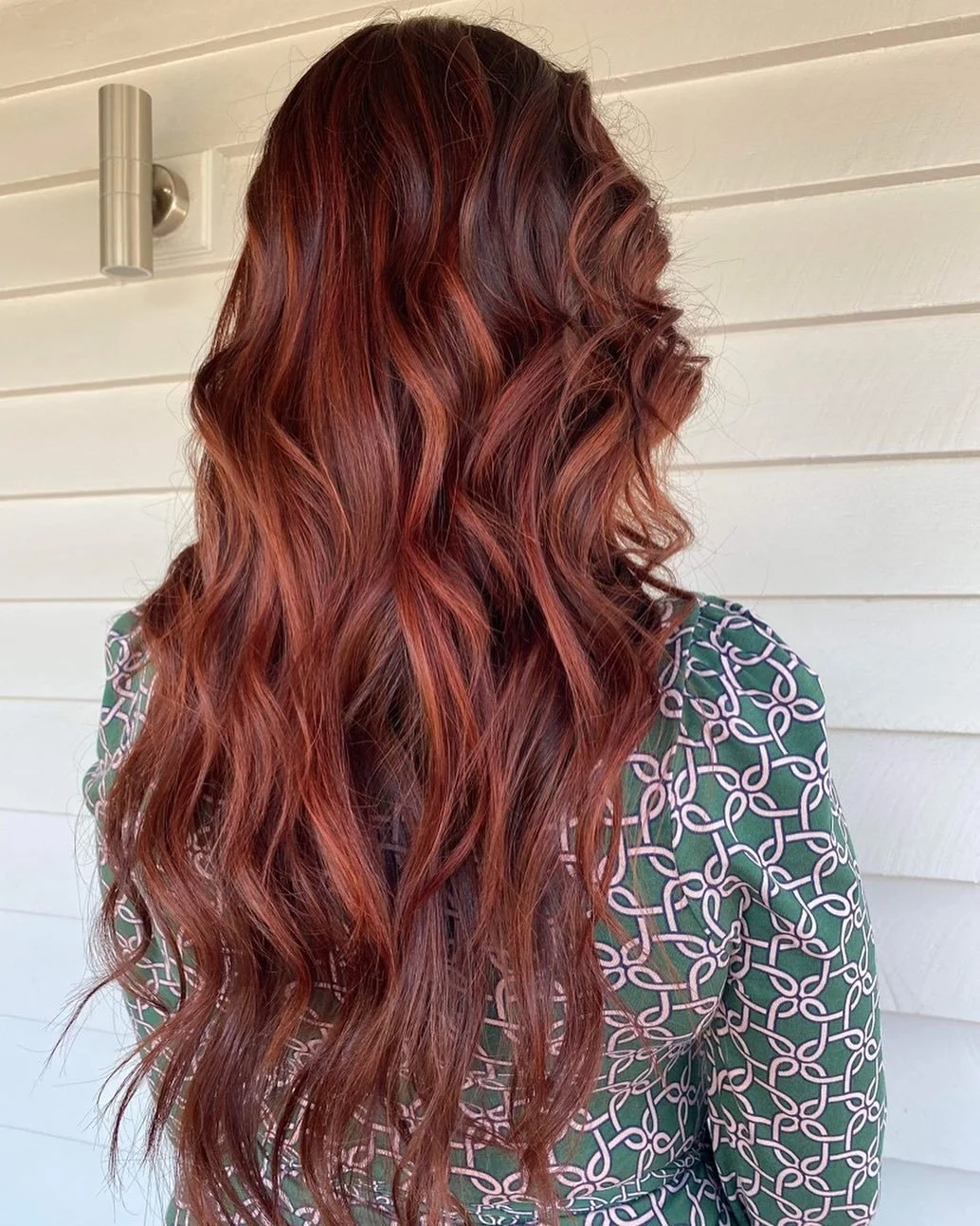The image shows a woman with long, wavy red hair standing against a white wooden wall, wearing a patterned green shirt.