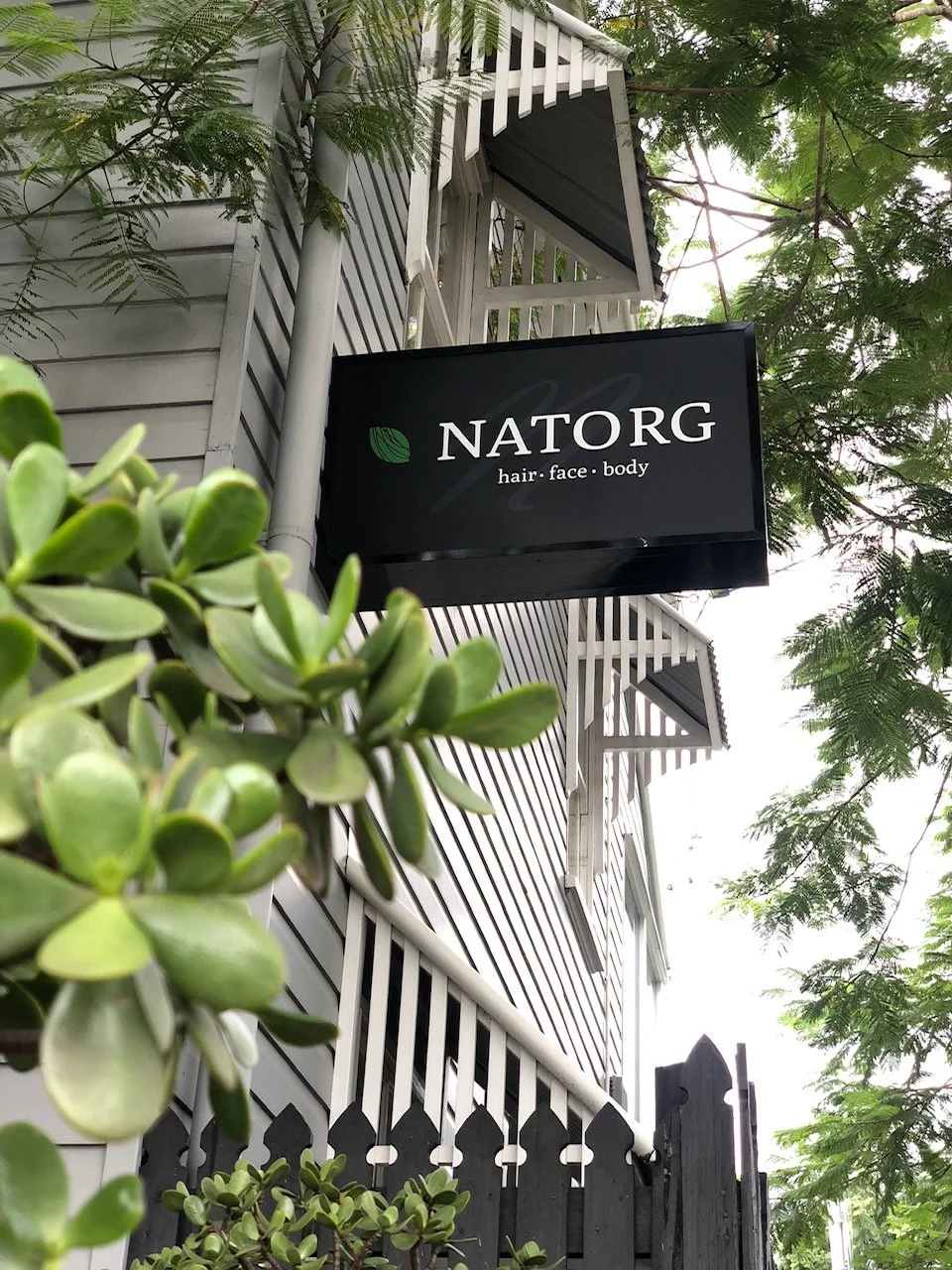 A black sign with the text 'NATORG' and 'hair face body' hangs outside a white wooden building with balconies, surrounded by green trees and plants.