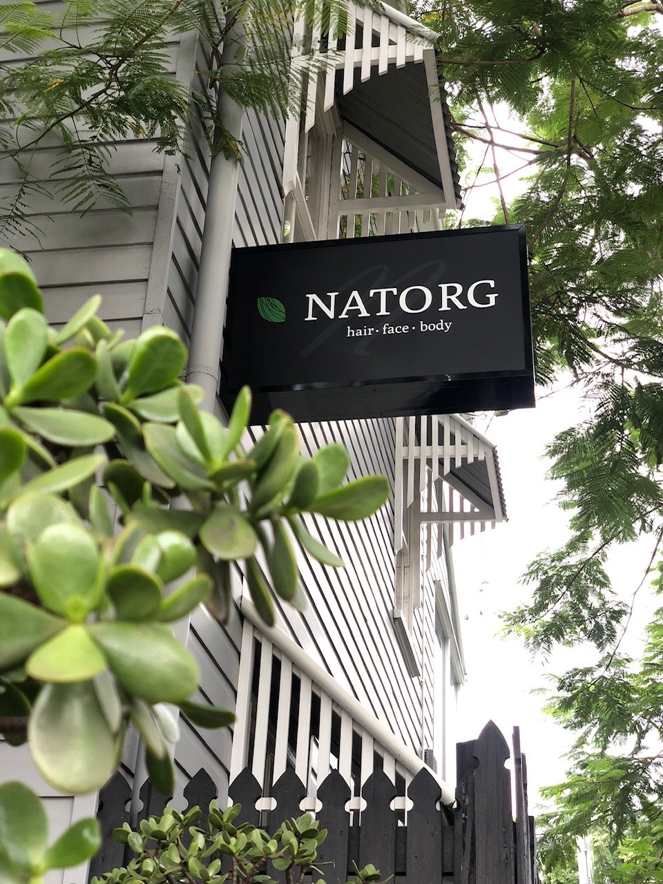 Outdoor sign for NATORG hair, face, and body salon, mounted on the building with greenery in the foreground and trees around.