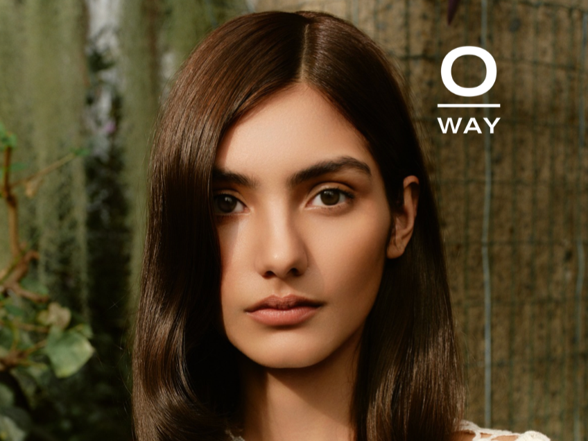A woman with long, straight brown hair looking directly at the camera in an outdoor setting with greenery in the background.