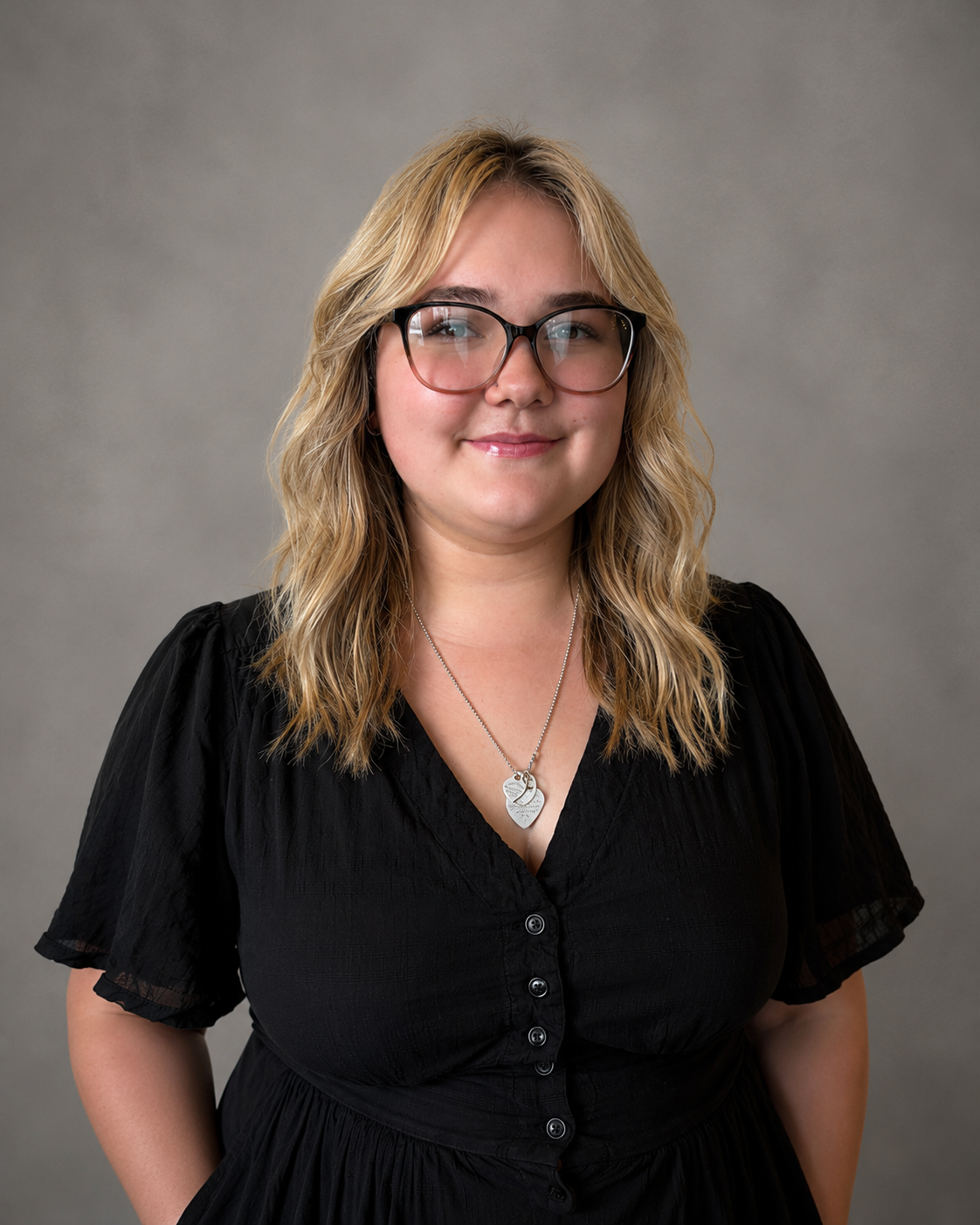 A woman with blonde wavy hair, glasses, wearing a black dress, and a heart-shaped necklace, standing against a gray background.