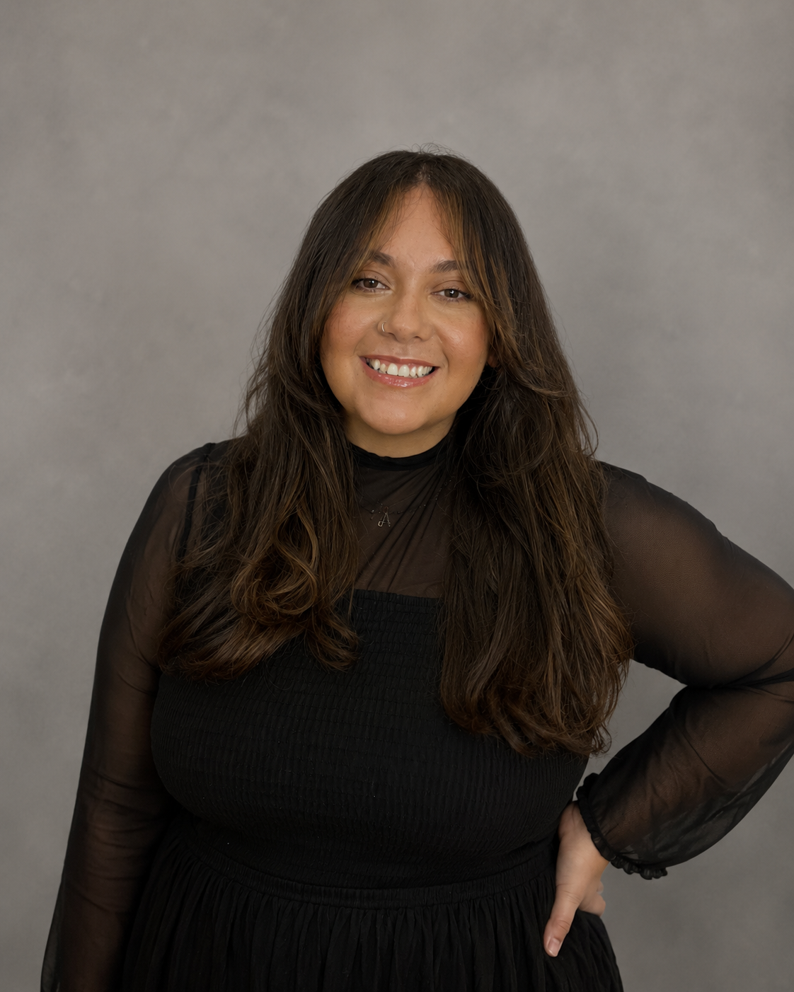 A young woman with long, wavy brown hair and a nose ring, smiling and wearing a black dress with sheer sleeves, standing against a gray background.