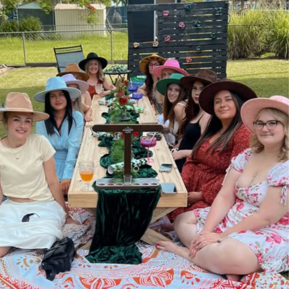 Group of women wearing various colorful wide-brimmed hats sitting around a decorated outdoor table in a backyard.
