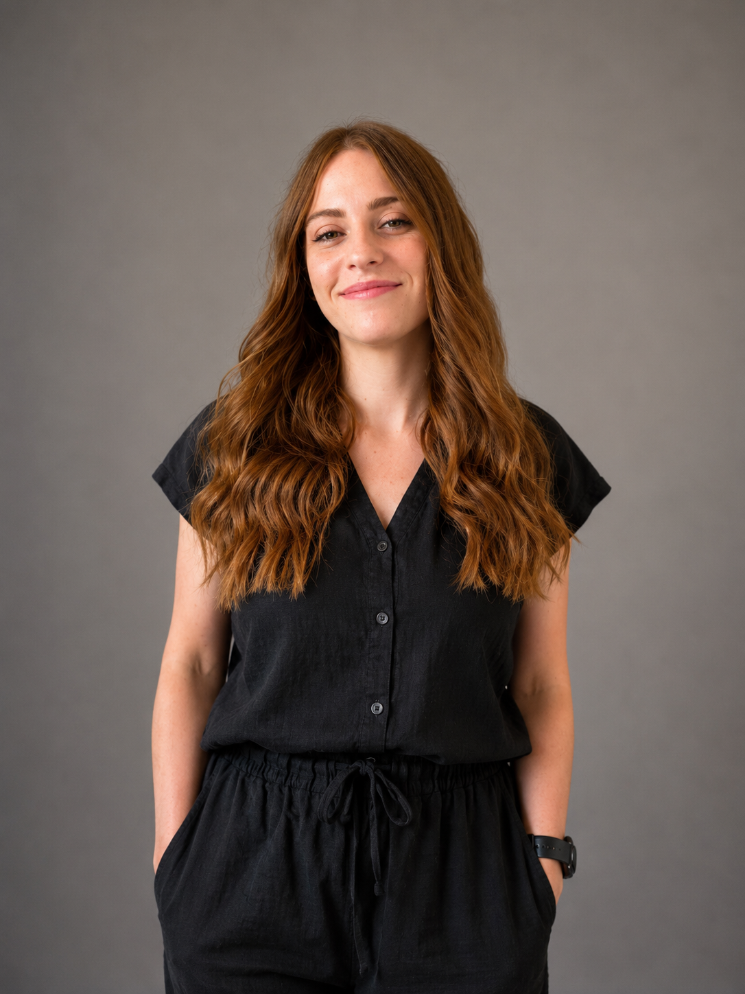 A young woman with long, wavy red hair wearing a black short-sleeve button-up top and matching black pants, smiling softly, standing against a plain gray background.